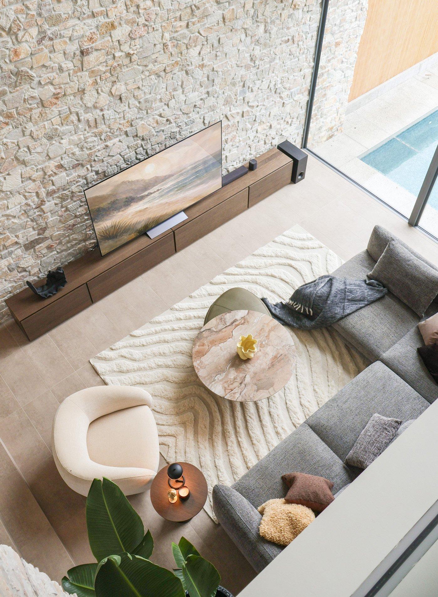 Moderne stue med grå sofa, beige stol, TV, og marmorert bord. Vegg i stein, stort vindu mot basseng.