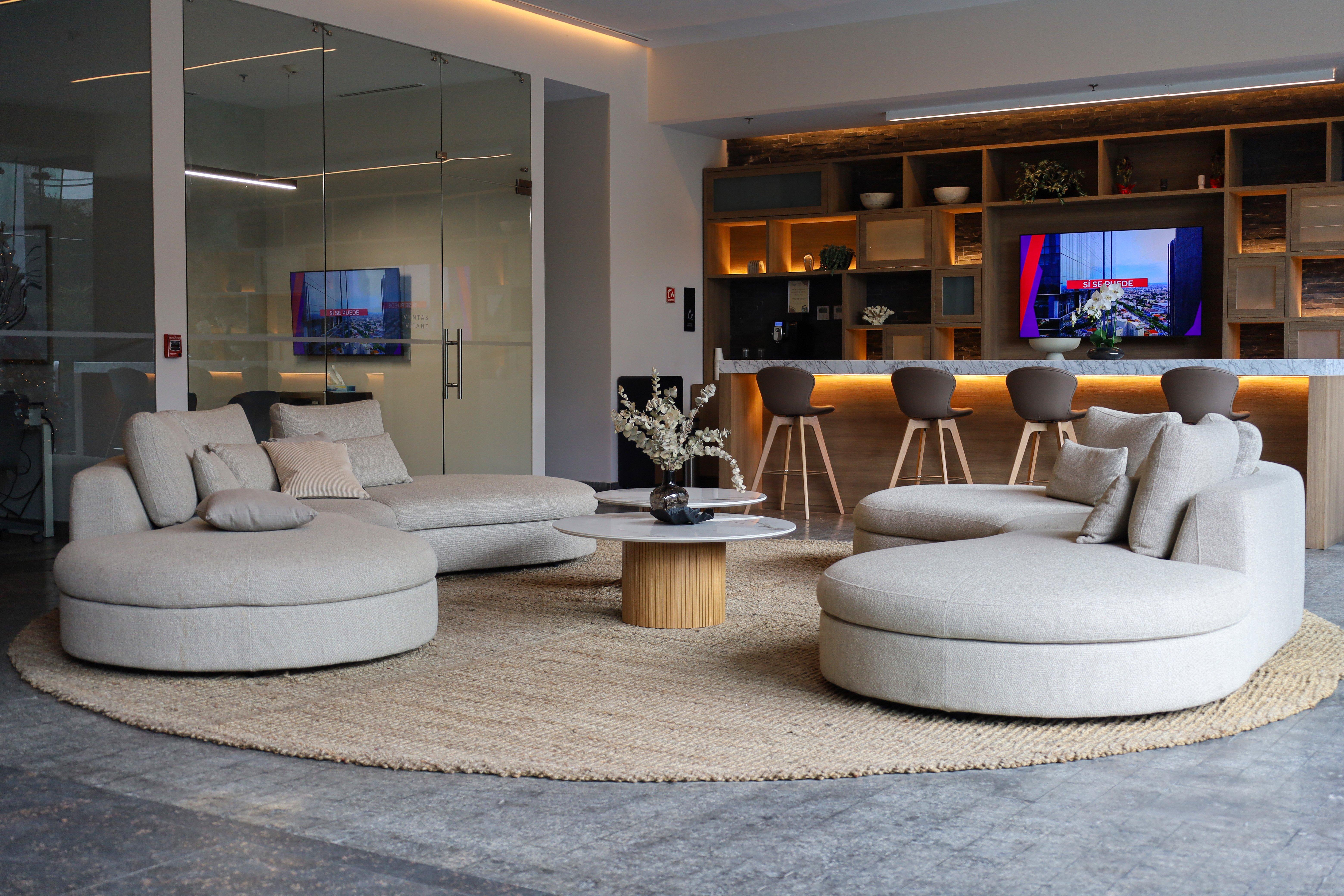 Modern lounge with round beige sofas, circular rug, and bar stools. TV on wall, decorative plants visible.