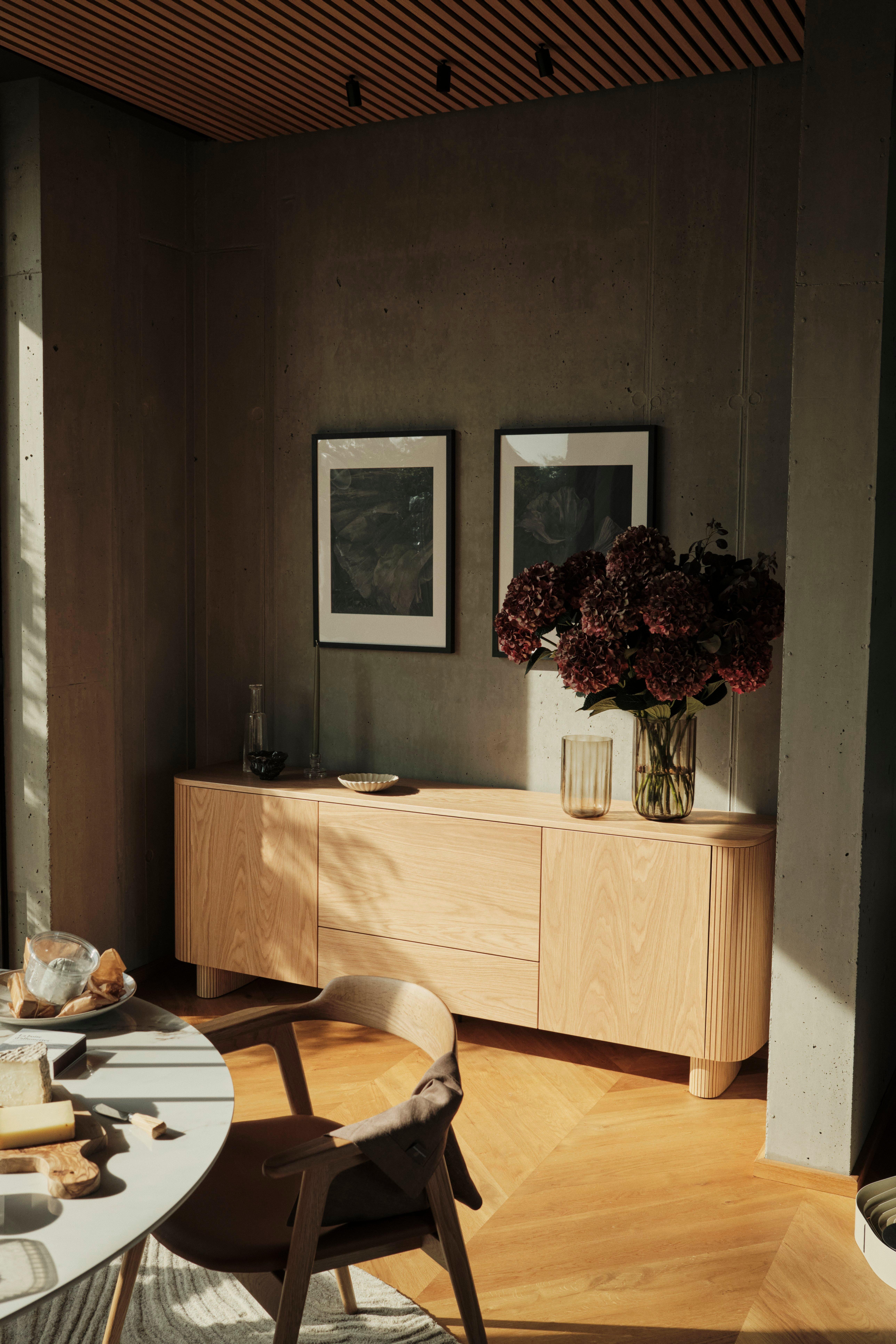 Moderný interiér so svetlým dreveným príborníkom, sušenými kvetmi vo váze, obrazom v ráme a čiastočne viditeľným jedálenským stolom v popredí.