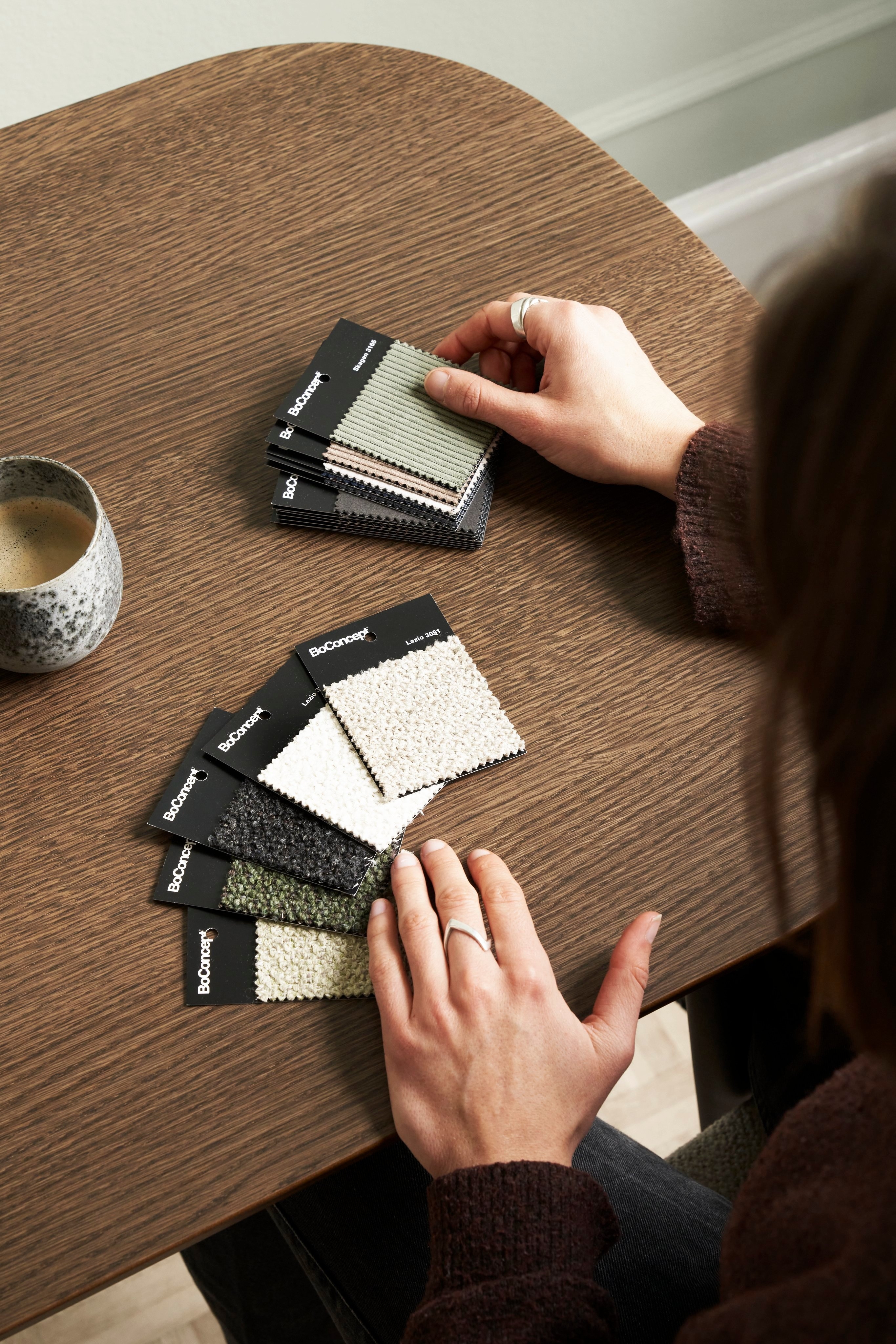 Person examining BoConcept fabric samples on a wooden table, next to a ceramic cup.