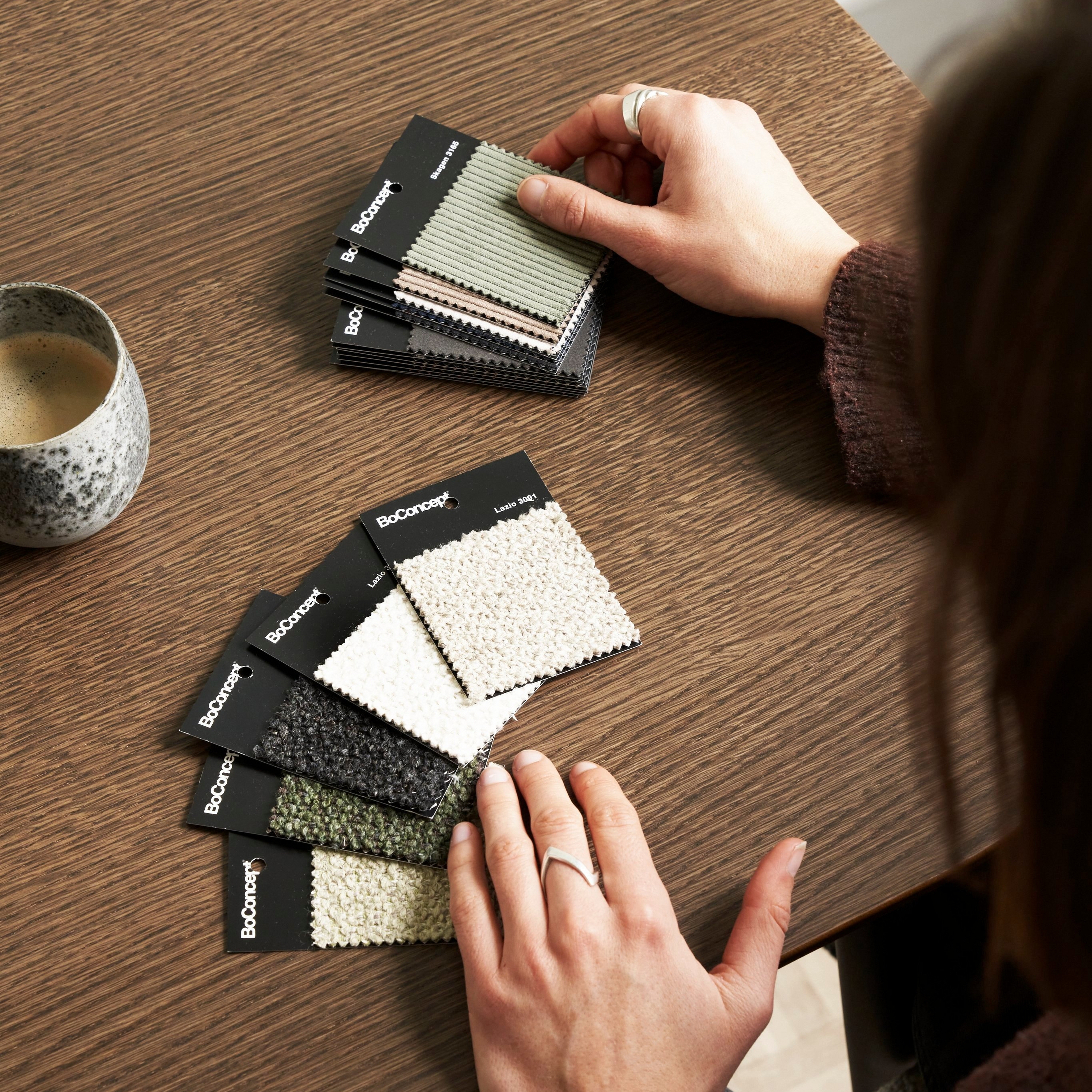 Person examining BoConcept fabric samples on a wooden table, next to a ceramic cup
