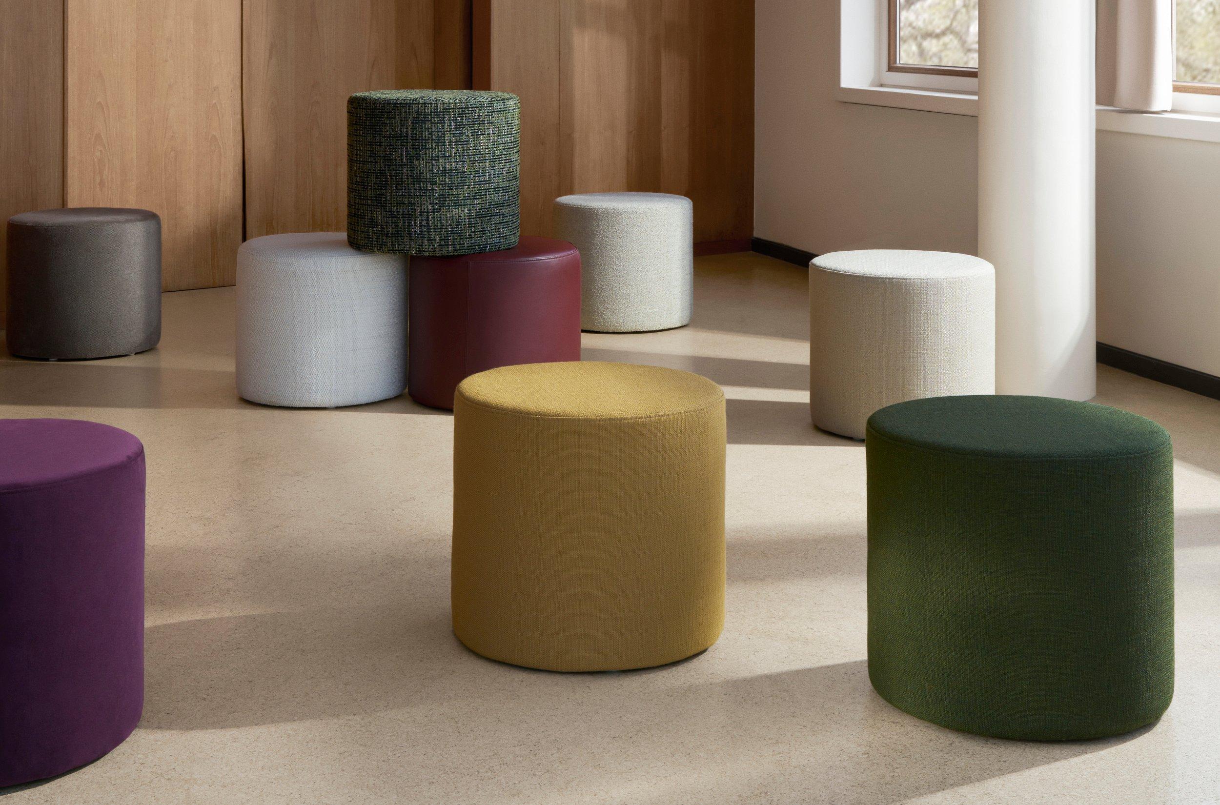 Assorted poufs in various shapes, colors, and textures arranged on wooden floor beside modern staircase and dark paneled wall.