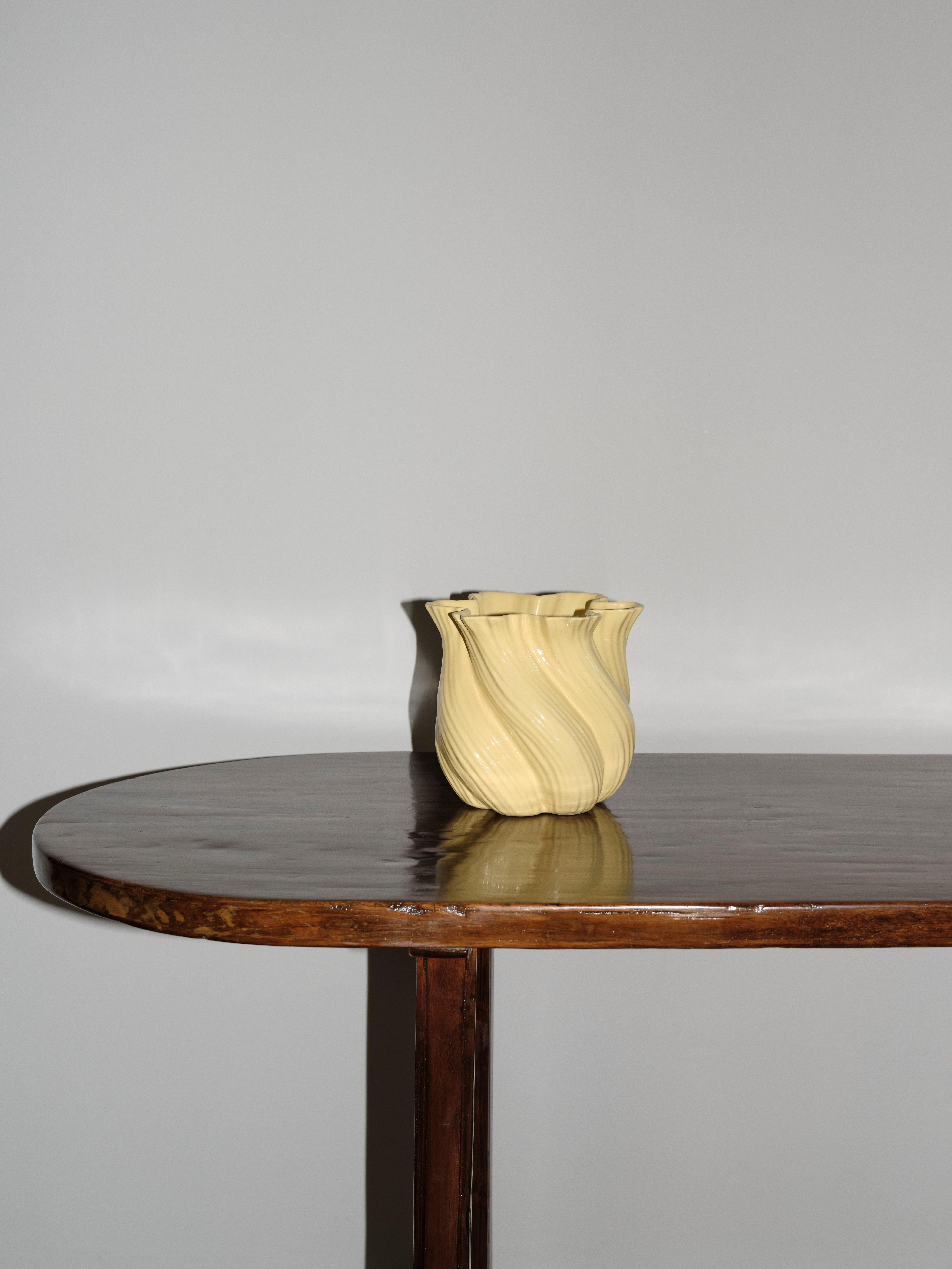 Buet gul keramisk vase med bølget tekstur plassert på et rundt bord i mørkt tre mot en ensfarget grå vegg.