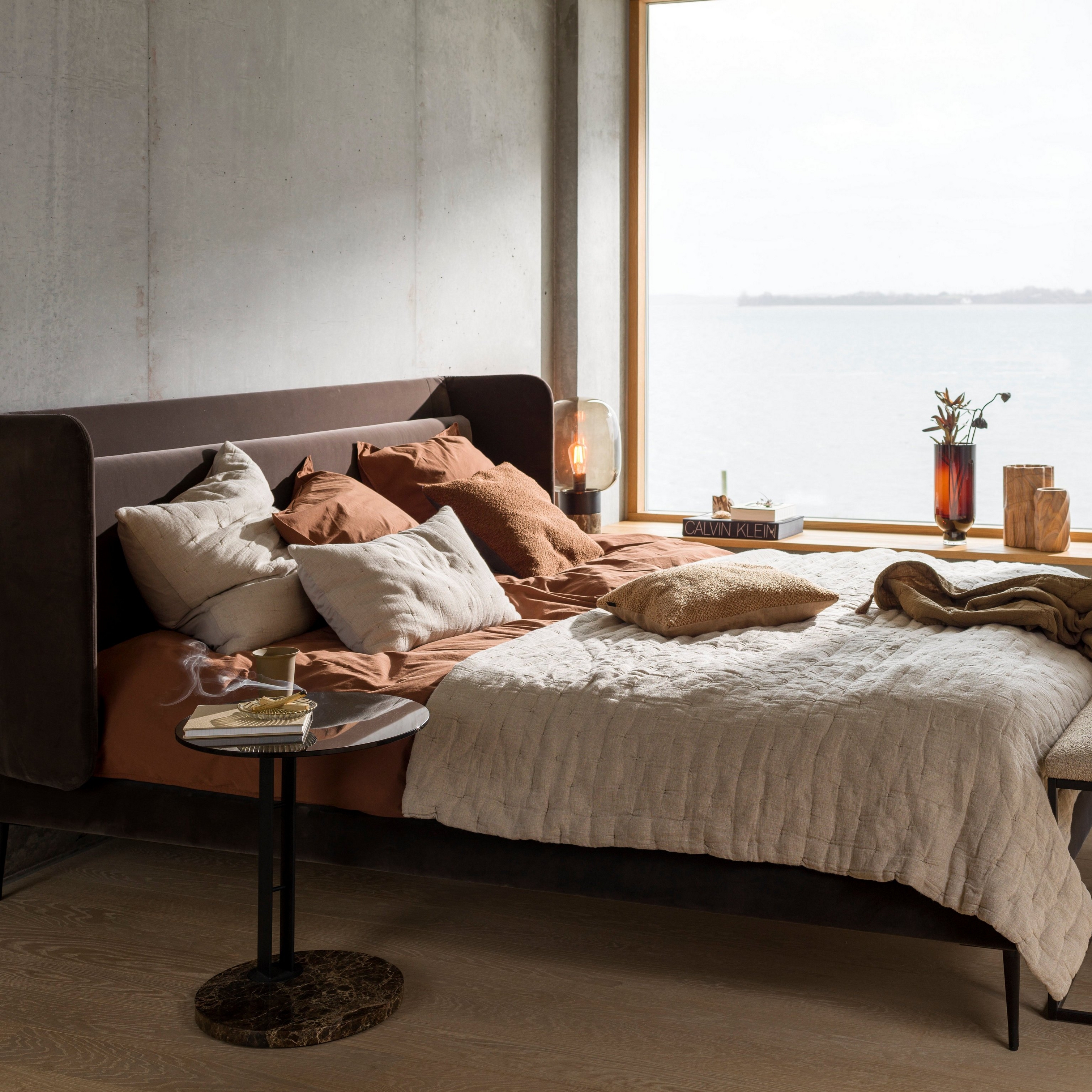 Mysigt sovrum med utsikt över vattnet, med sängkläder i jordnära toner och ett litet sidobord.
