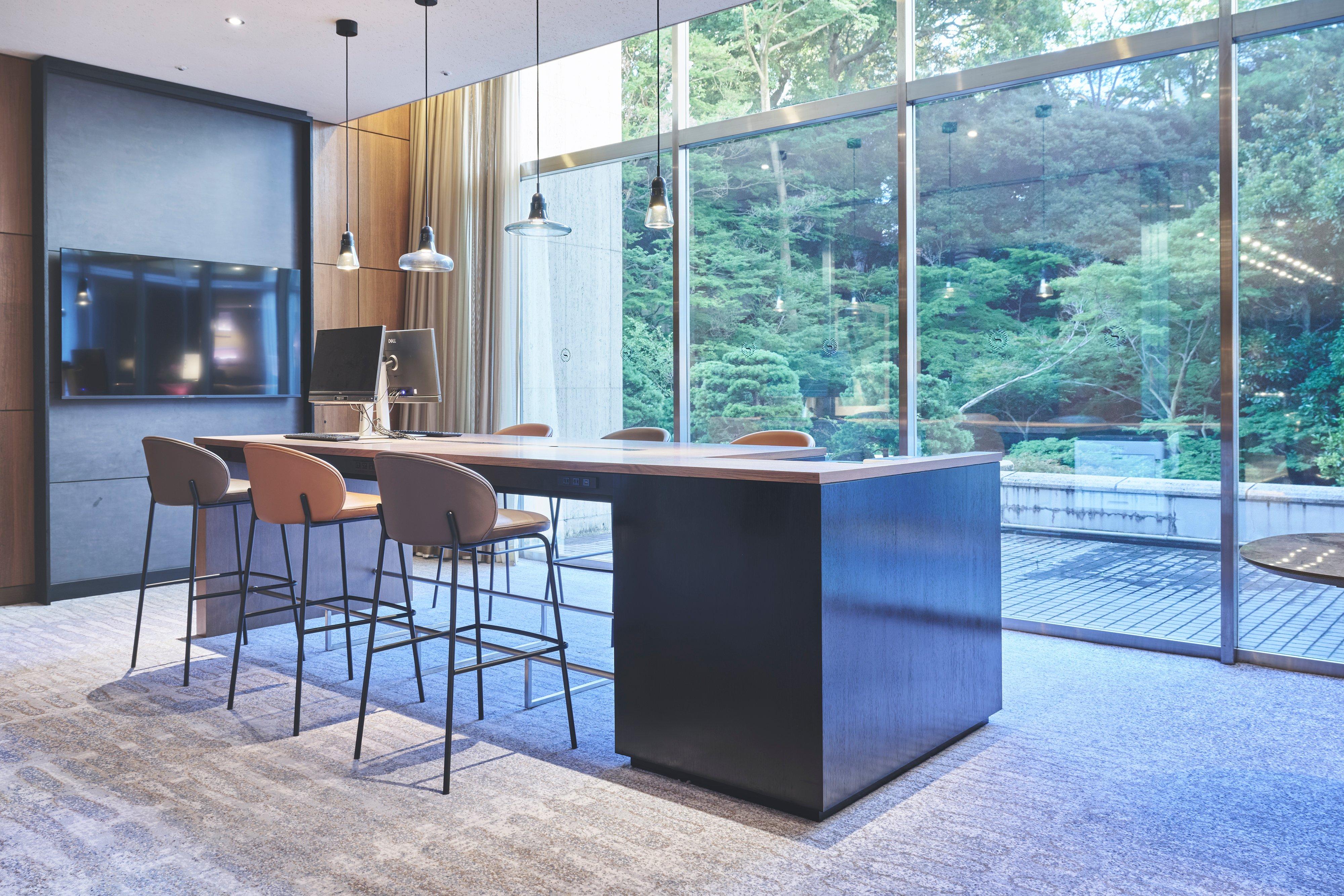 Modern communal table with bar stools under pendant lights, in a glass-walled lounge area.