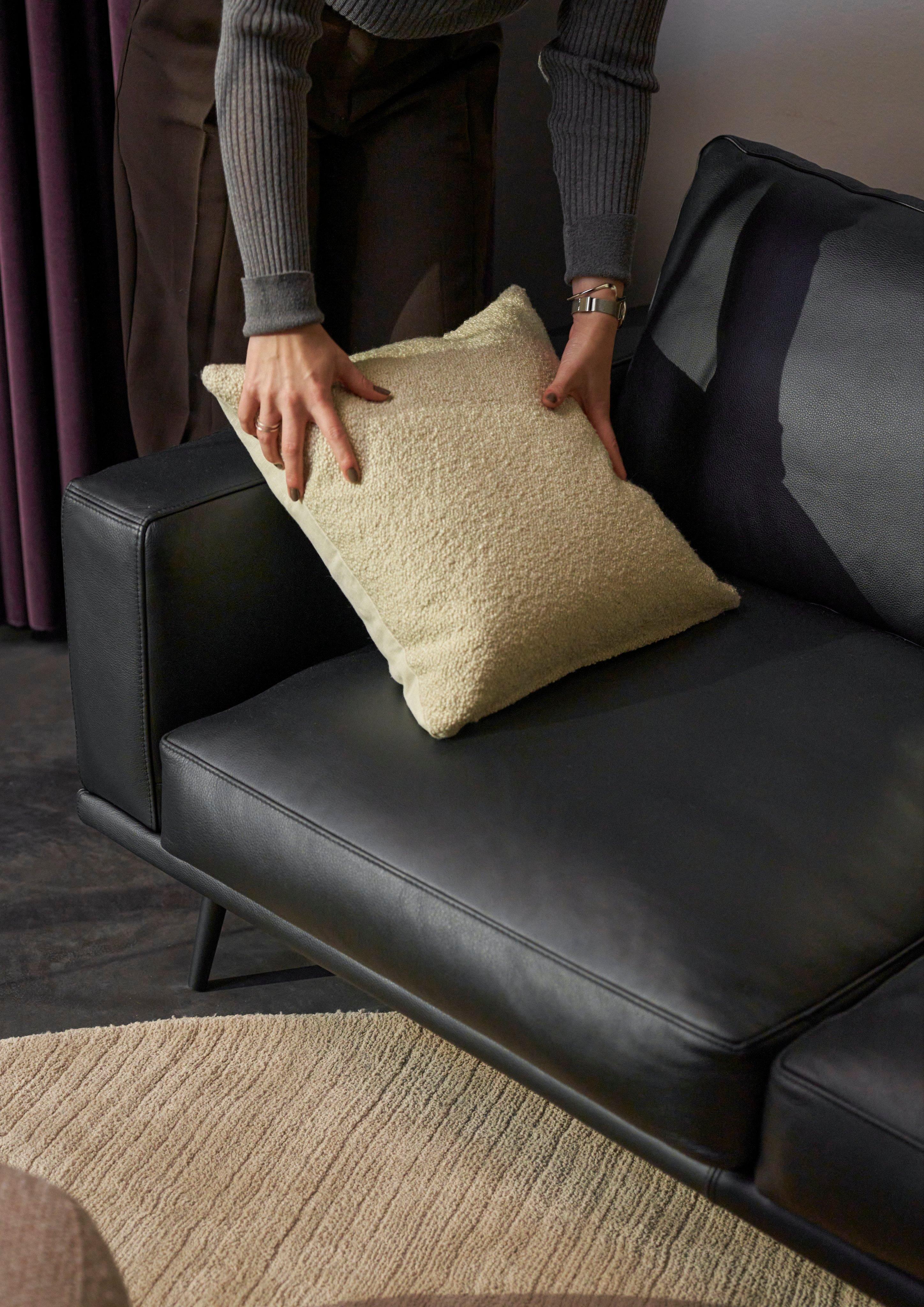 Person placing a cream bouclé cushion on a black Nordic Grain leather Carlton sofa, oat-colored Fiona rug below.