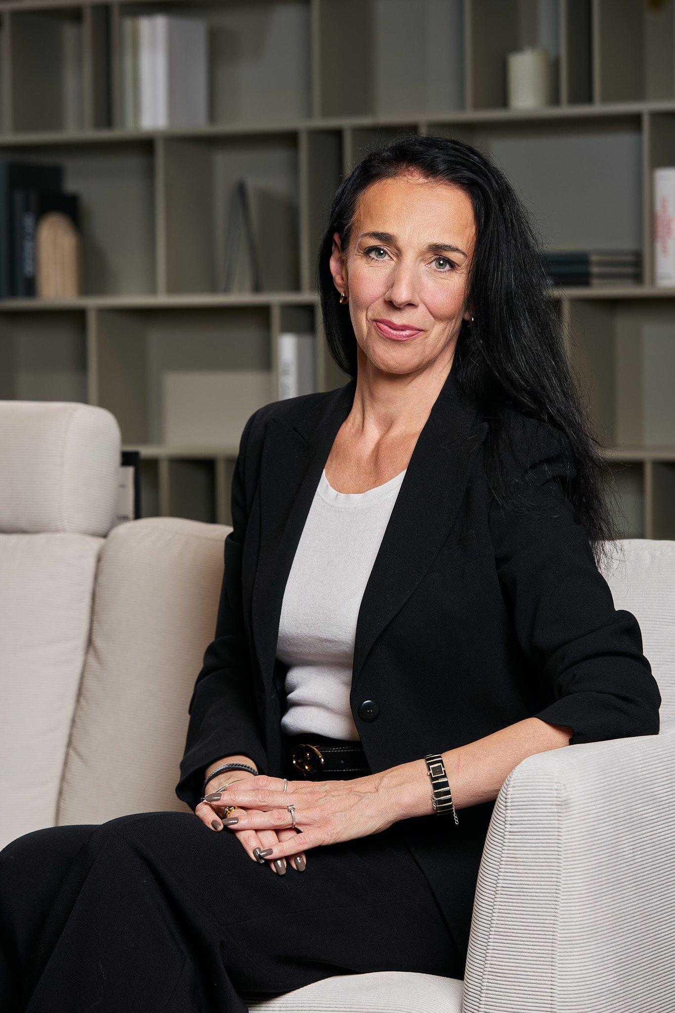 Žena v černém saku sedí na pohovce a dívá se do kamery.