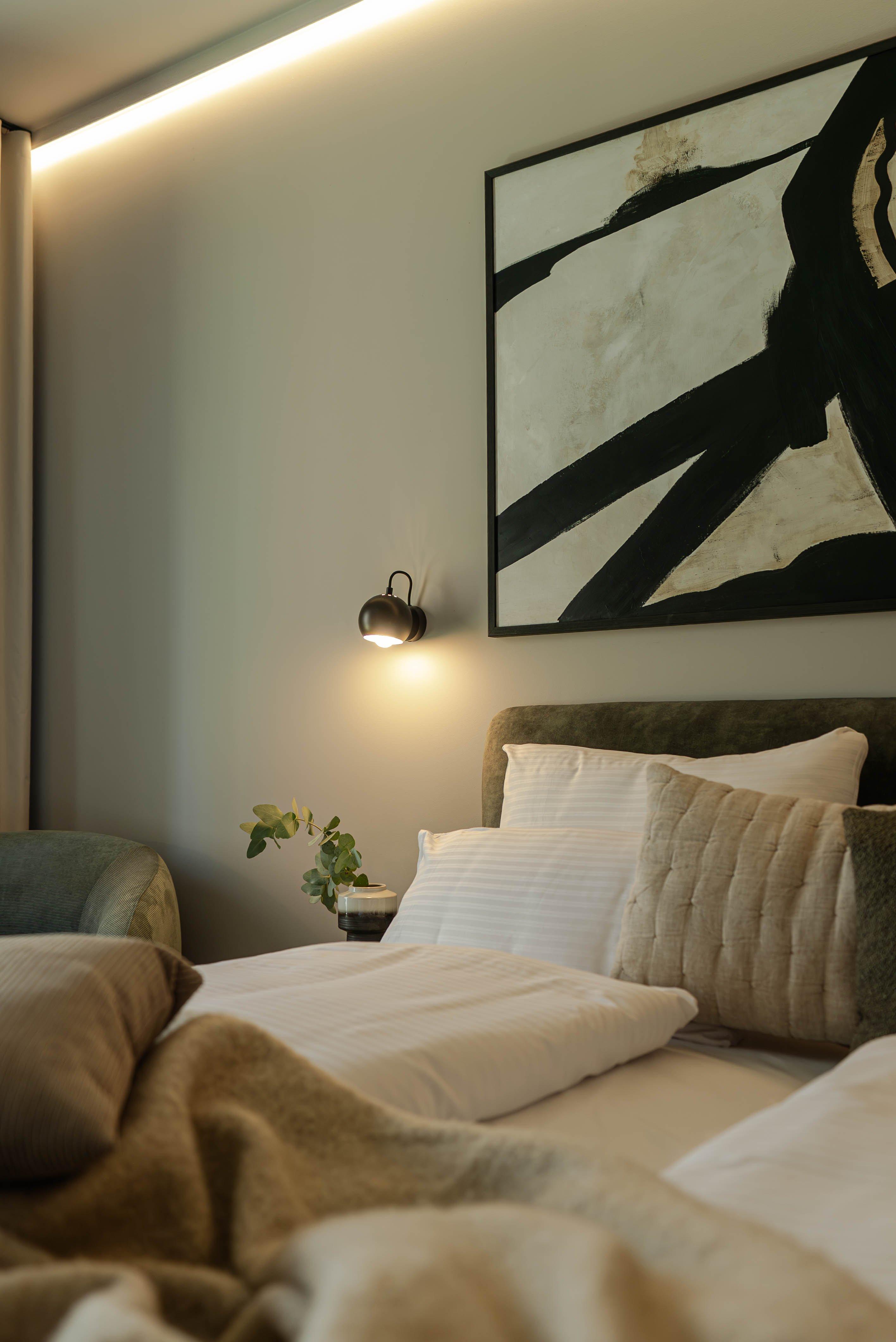 Lit avec oreillers, plaid, lampe murale et tableau abstrait. Plante verte sur la table.
