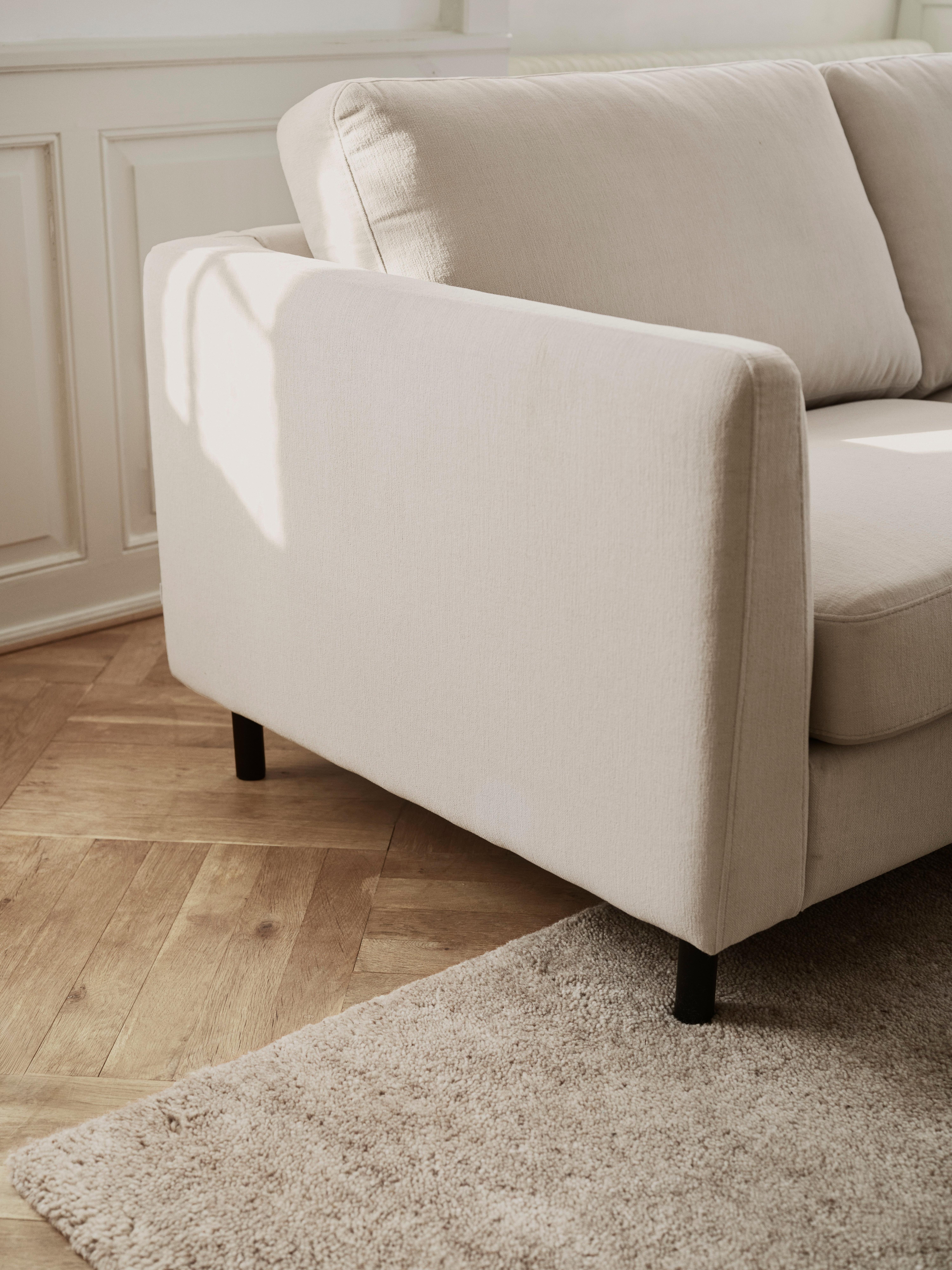 Sofá de tela verde oliva con un cojín acanalado beige, colocado cerca de grandes ventanales de cristal y una alfombra blanca texturizada.