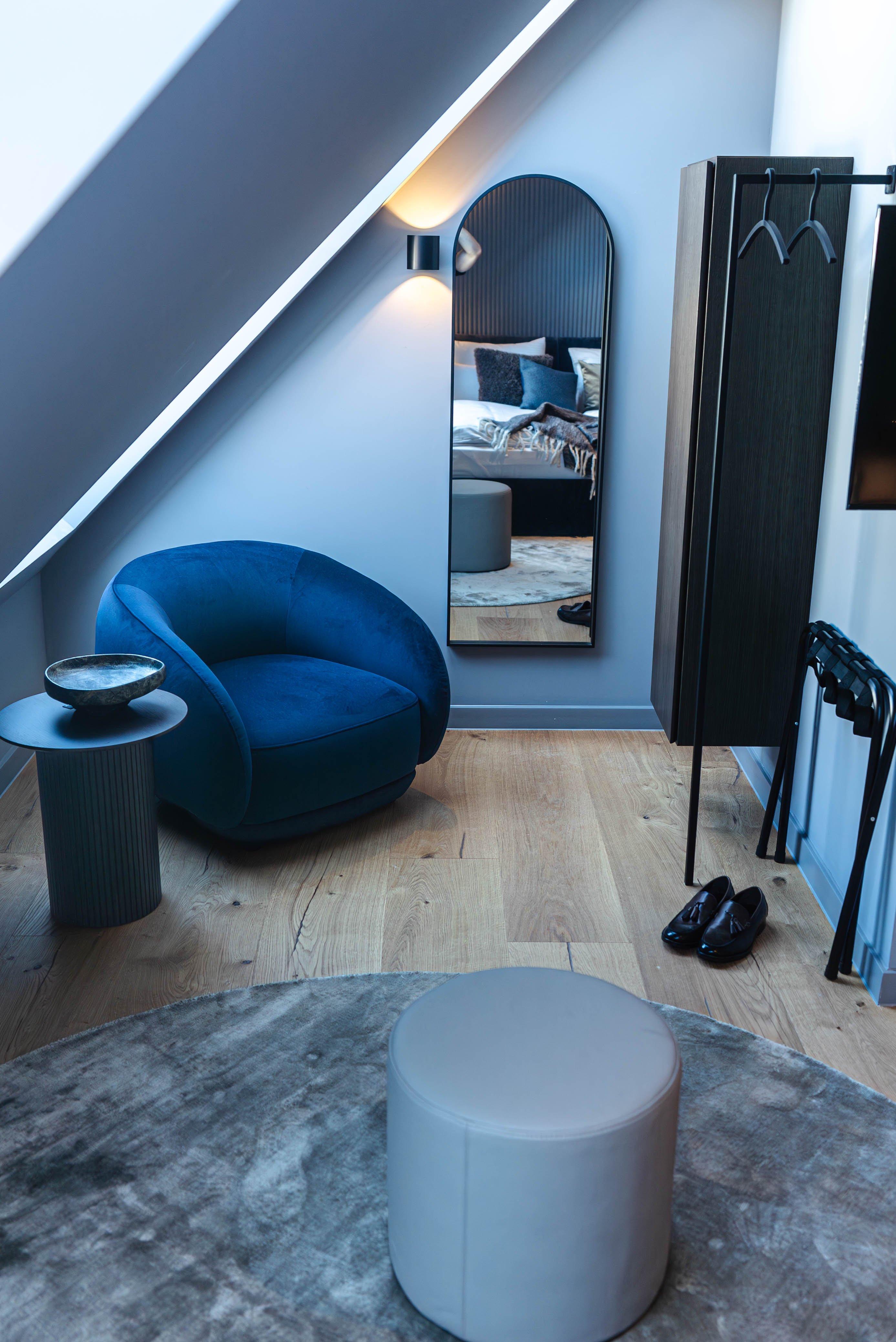 Fauteuil bleu, miroir, chaussures, et pouf dans chambre mansardée.