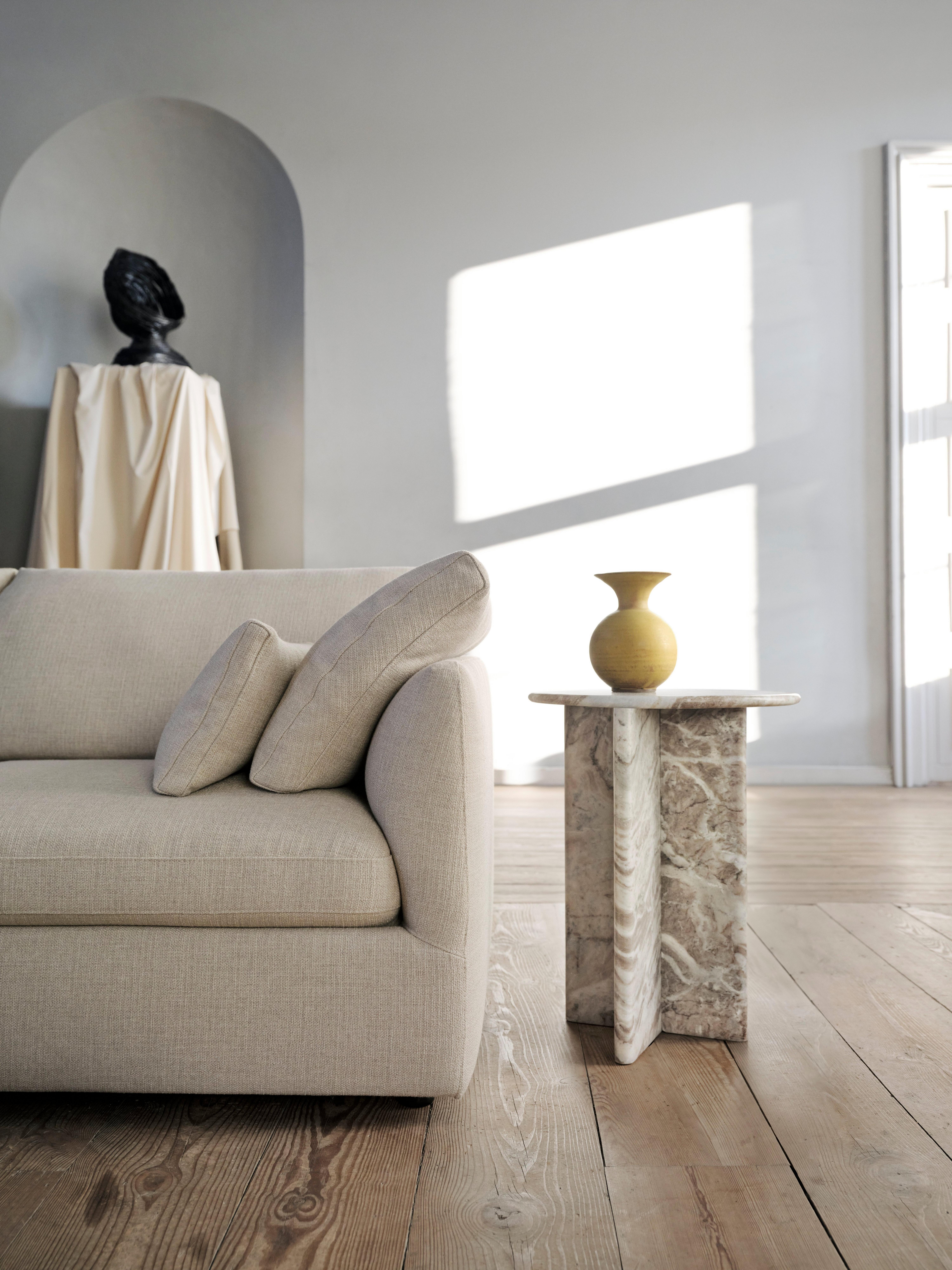 Beige kangassohva ja kaksi tyynyä, marmorinen sivupöytä ja pyöreä koriste-esine puulattialla valoisassa huoneessa.