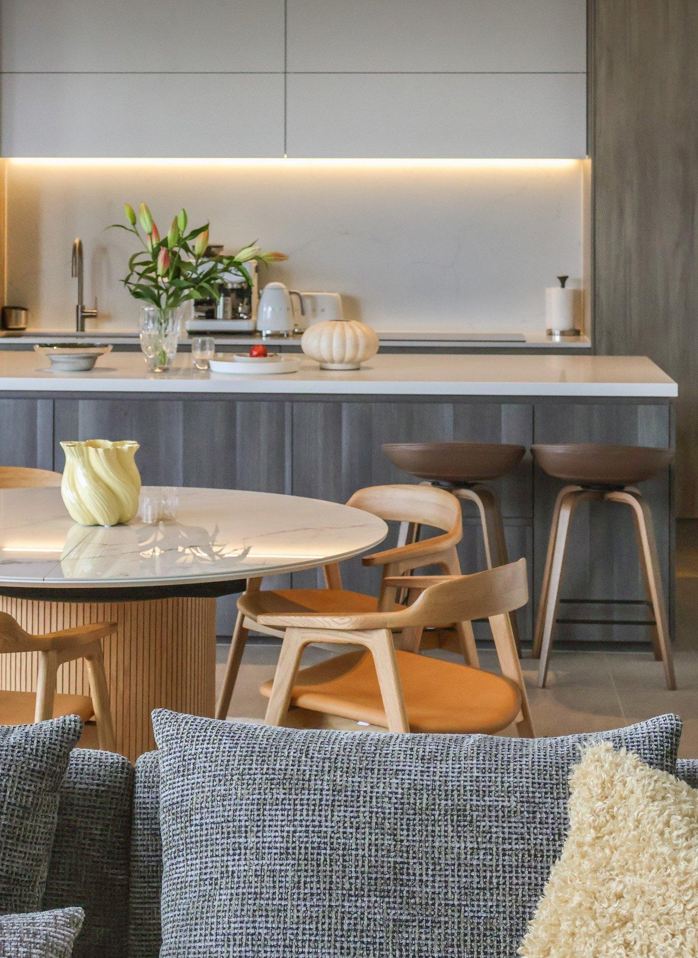 Moderne kjøkken med øy, blomster, stoler og bord. Brun barstol. Lys vase på bordet.