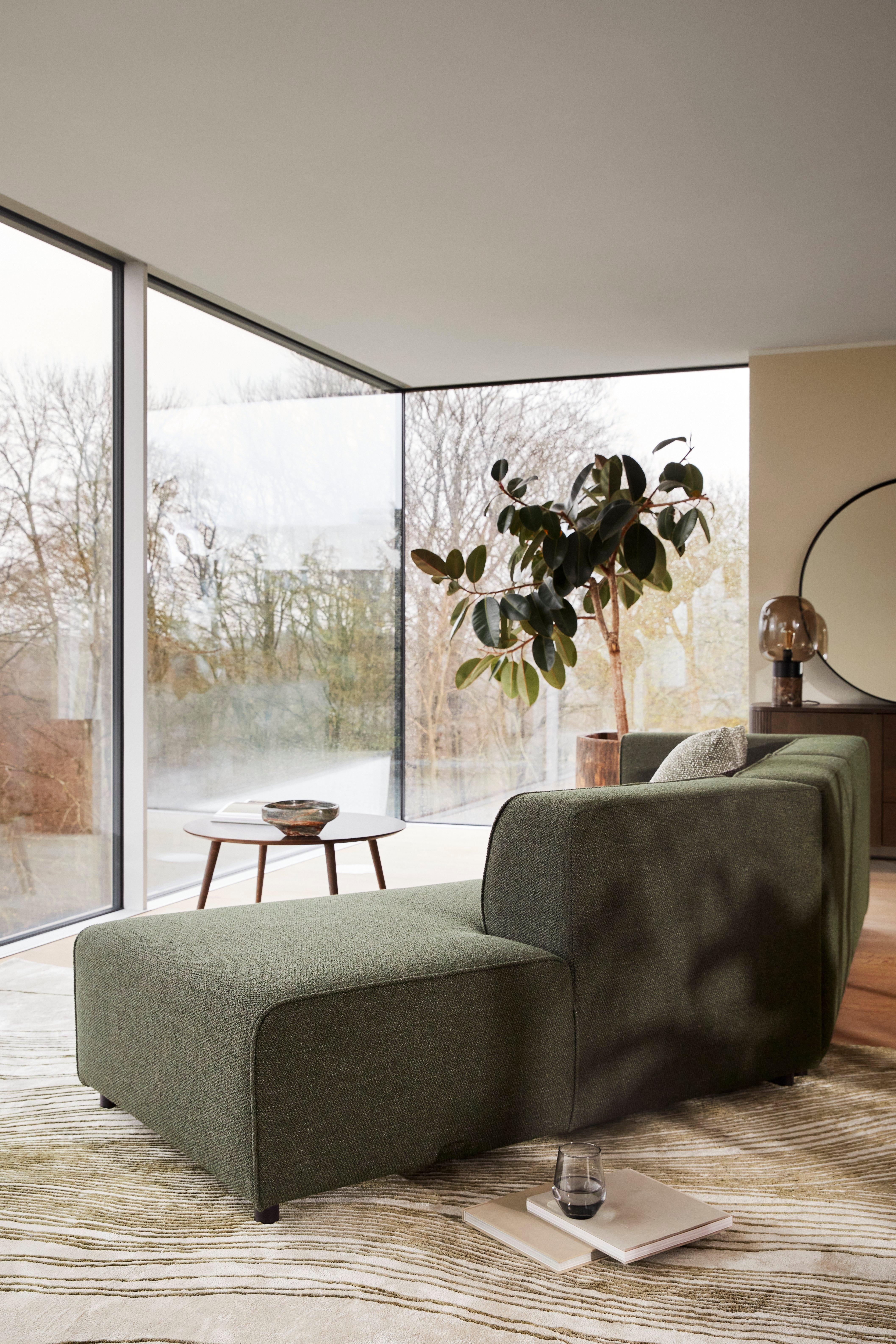 Pokój dzienny z zieloną sofą Carmo, okrągłym stolikiem kawowym Bornholm, rośliną doniczkową, dużymi oknami i teksturowanym dywanem.