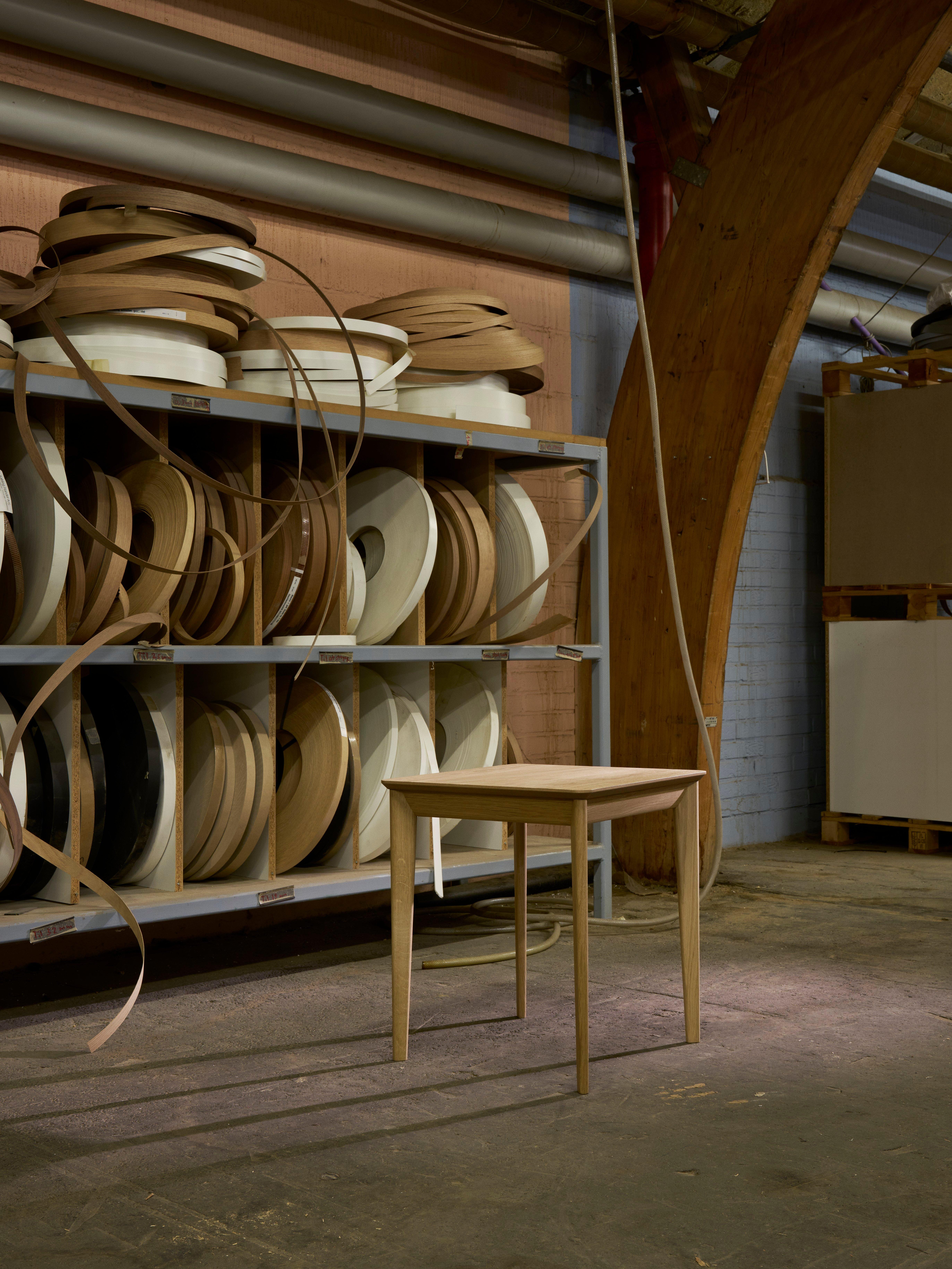Aarhus side table in natural oak veneer with tapered legs, placed in an industrial space with shelves of wood edging rolls.
