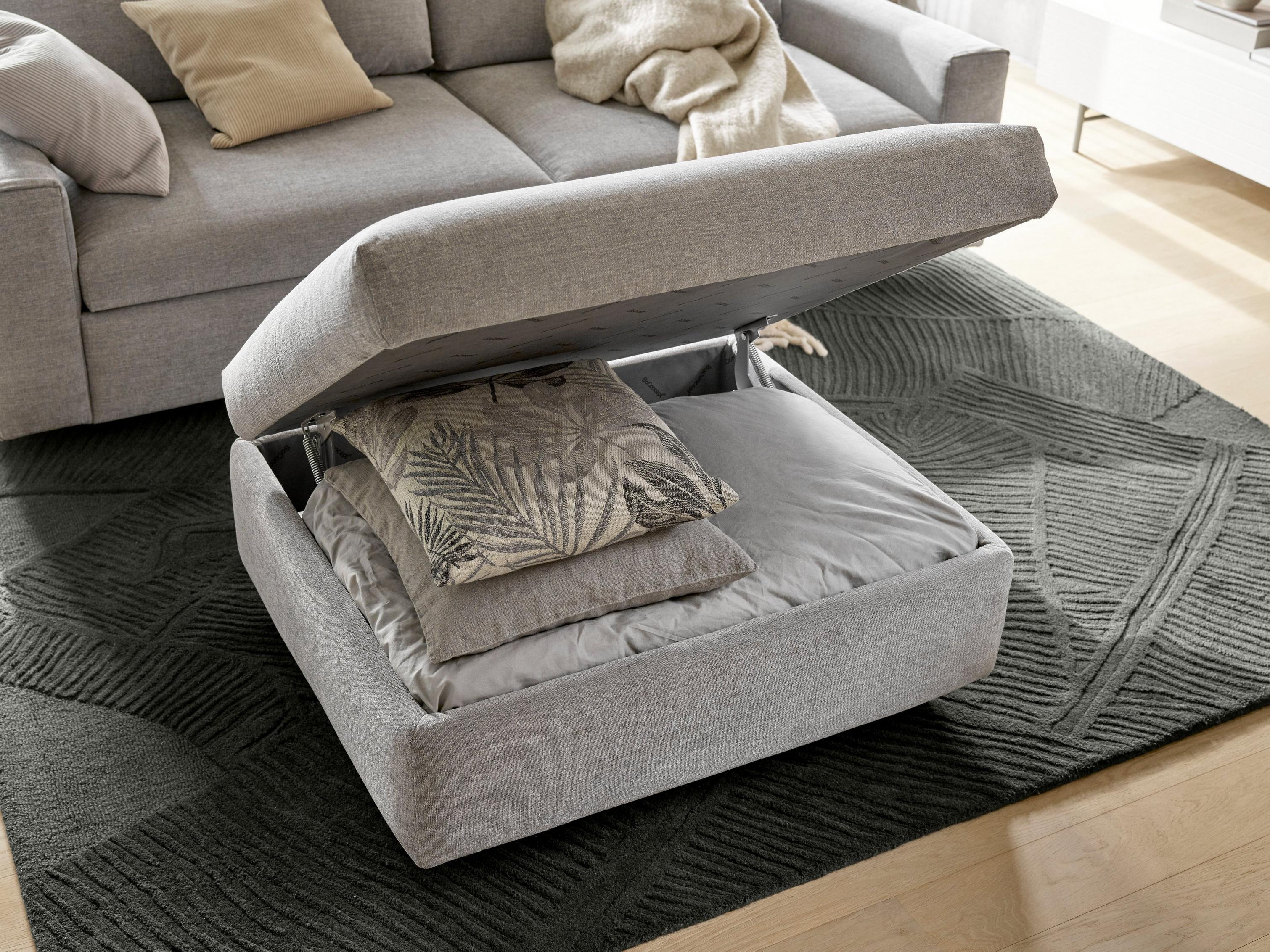 Gray upholstered footstool with storage, open showing pillows, by a sofa on a textured rug.