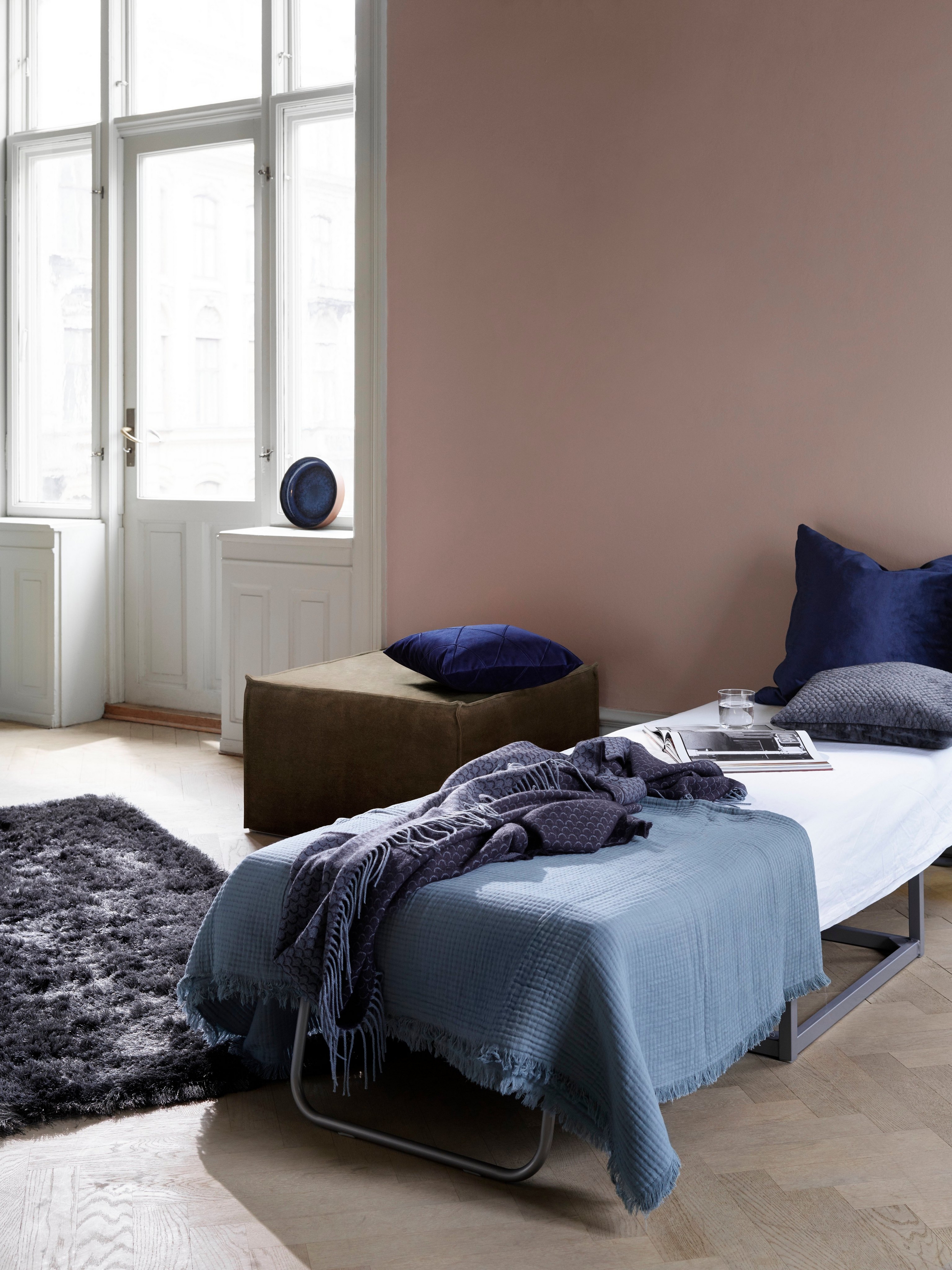 Indbydende værelse med gæsteseng, blåt sengetøj og mørkt, blødt tæppe tæt på oplyste vinduer og Xtra fodskammel.