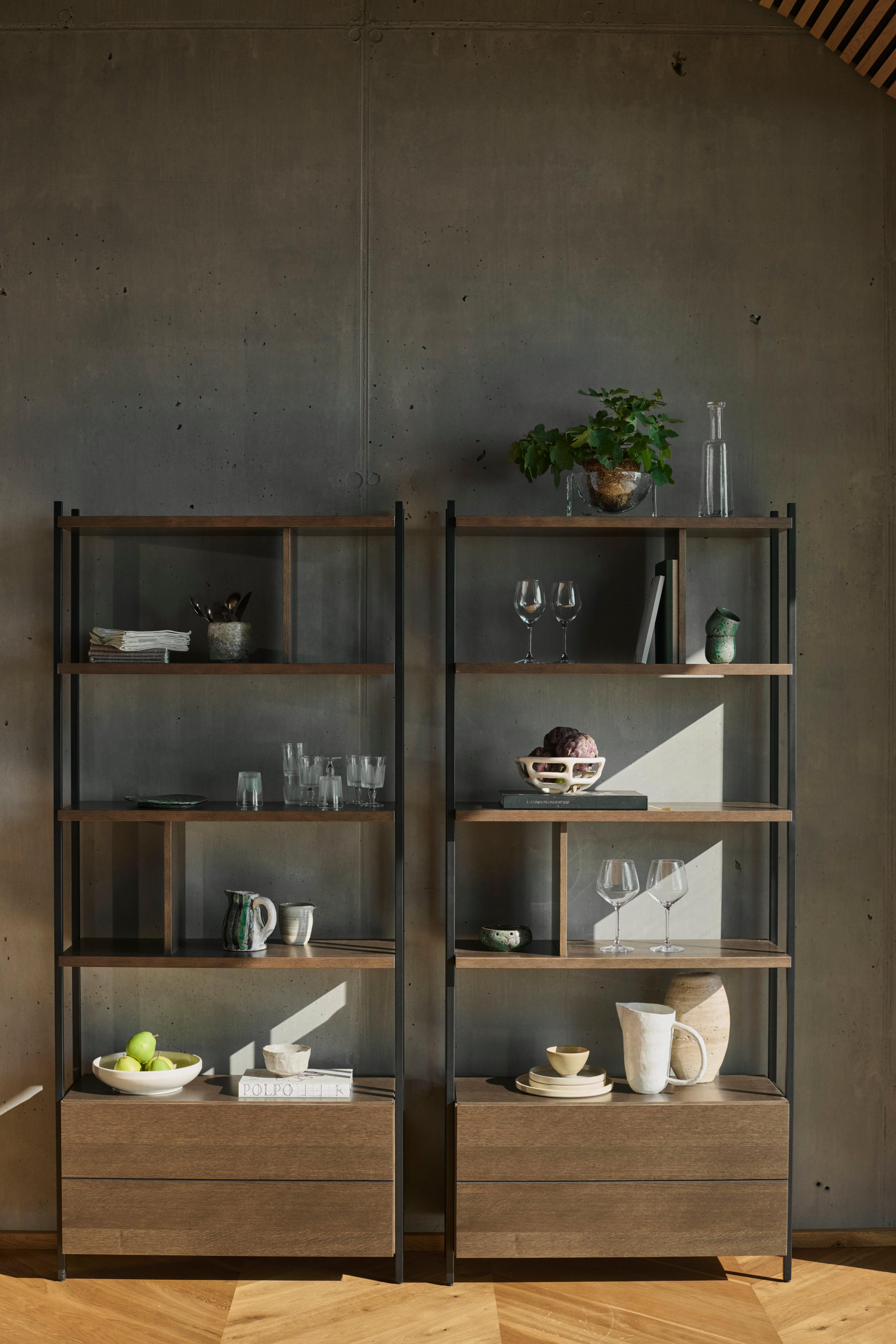 Two open shelving units with wood shelves and drawers, styled with glassware, ceramics, books, and a potted plant against a dark wall