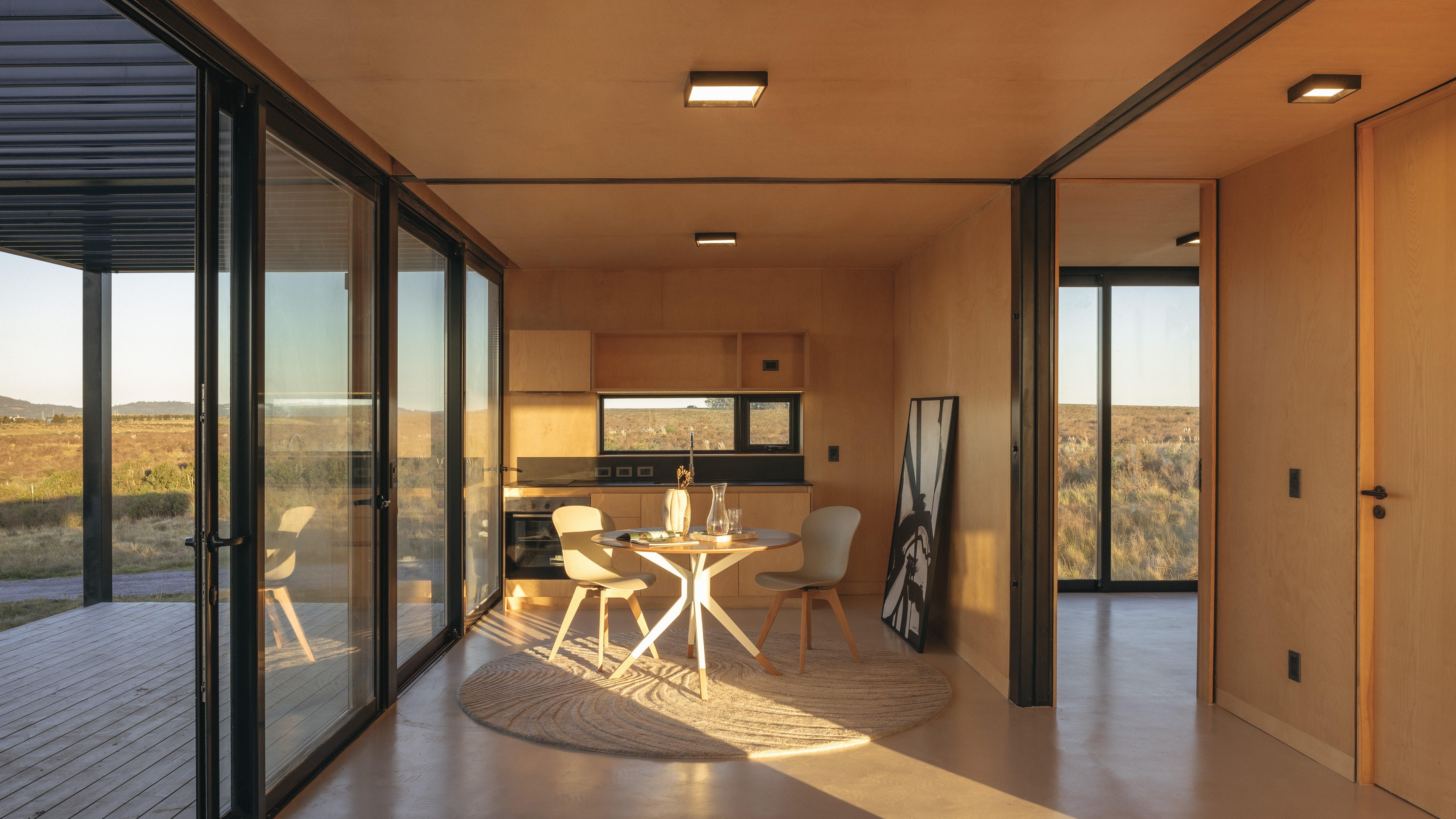 Modern interior with round table, two chairs, kitchen, and large windows overlooking a landscape.