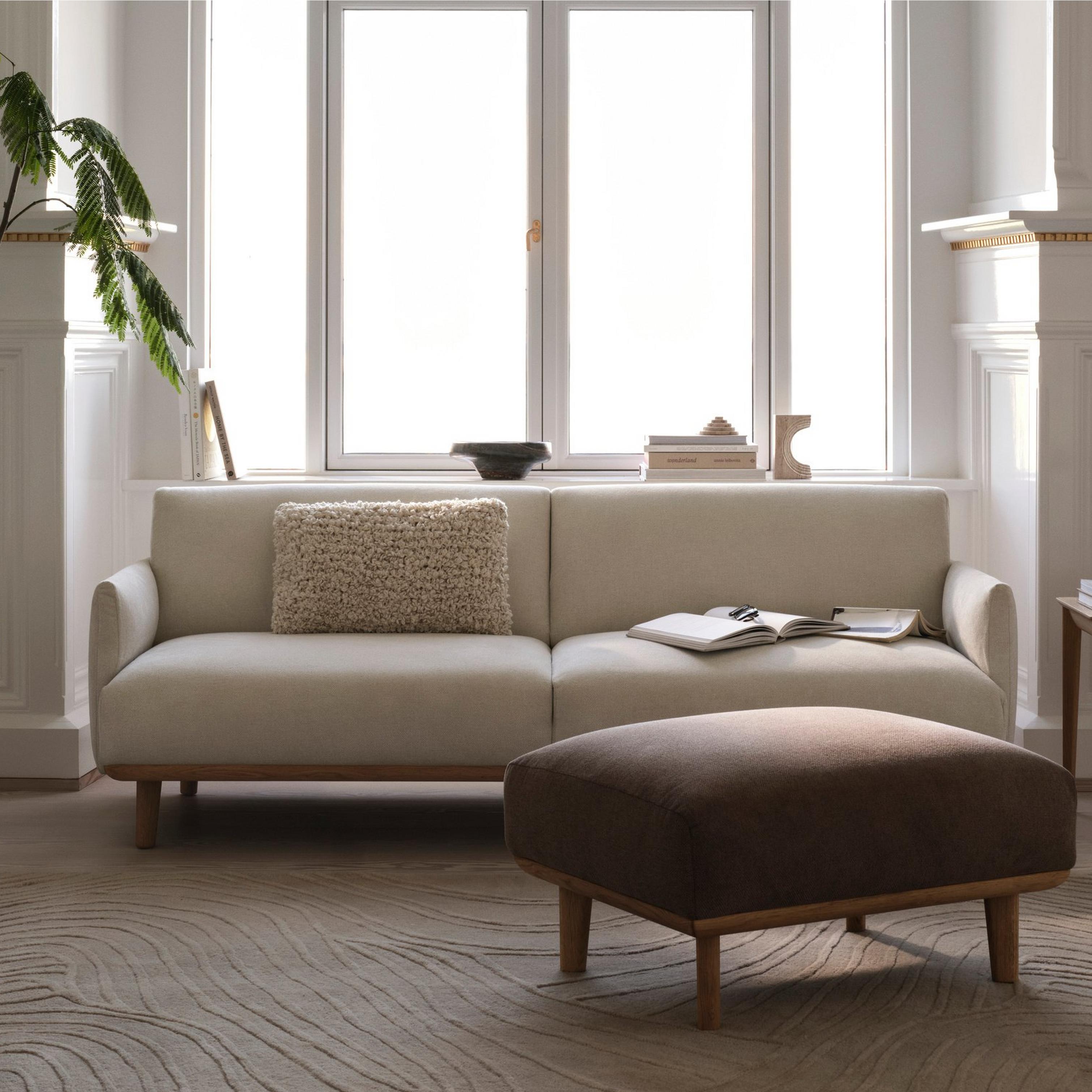 Elegant living room with cream sofa, brown ottoman, and large indoor plant by the window.