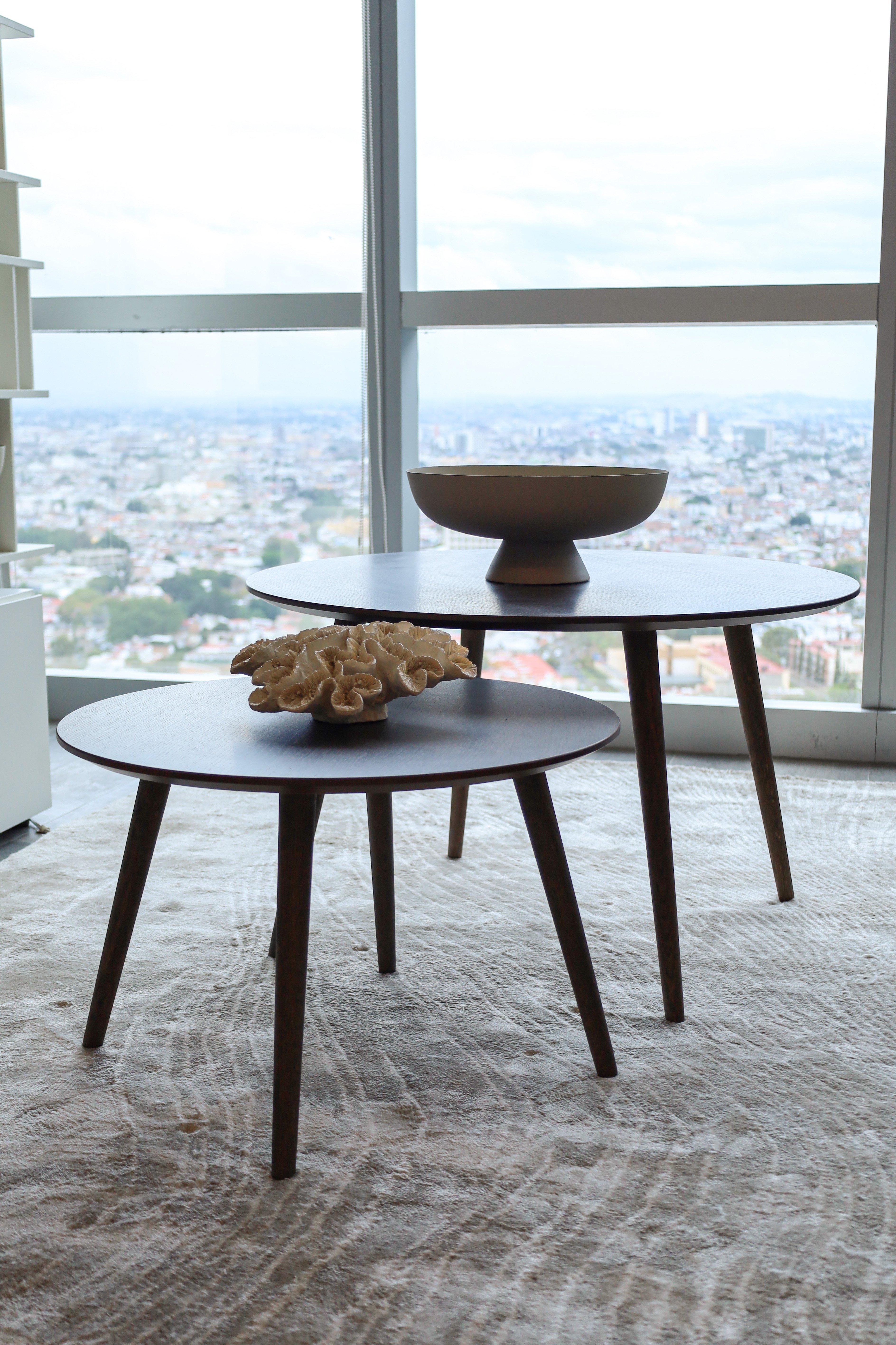 Two round wooden tables, one with a bowl, the other with a coral decoration. City view outside.