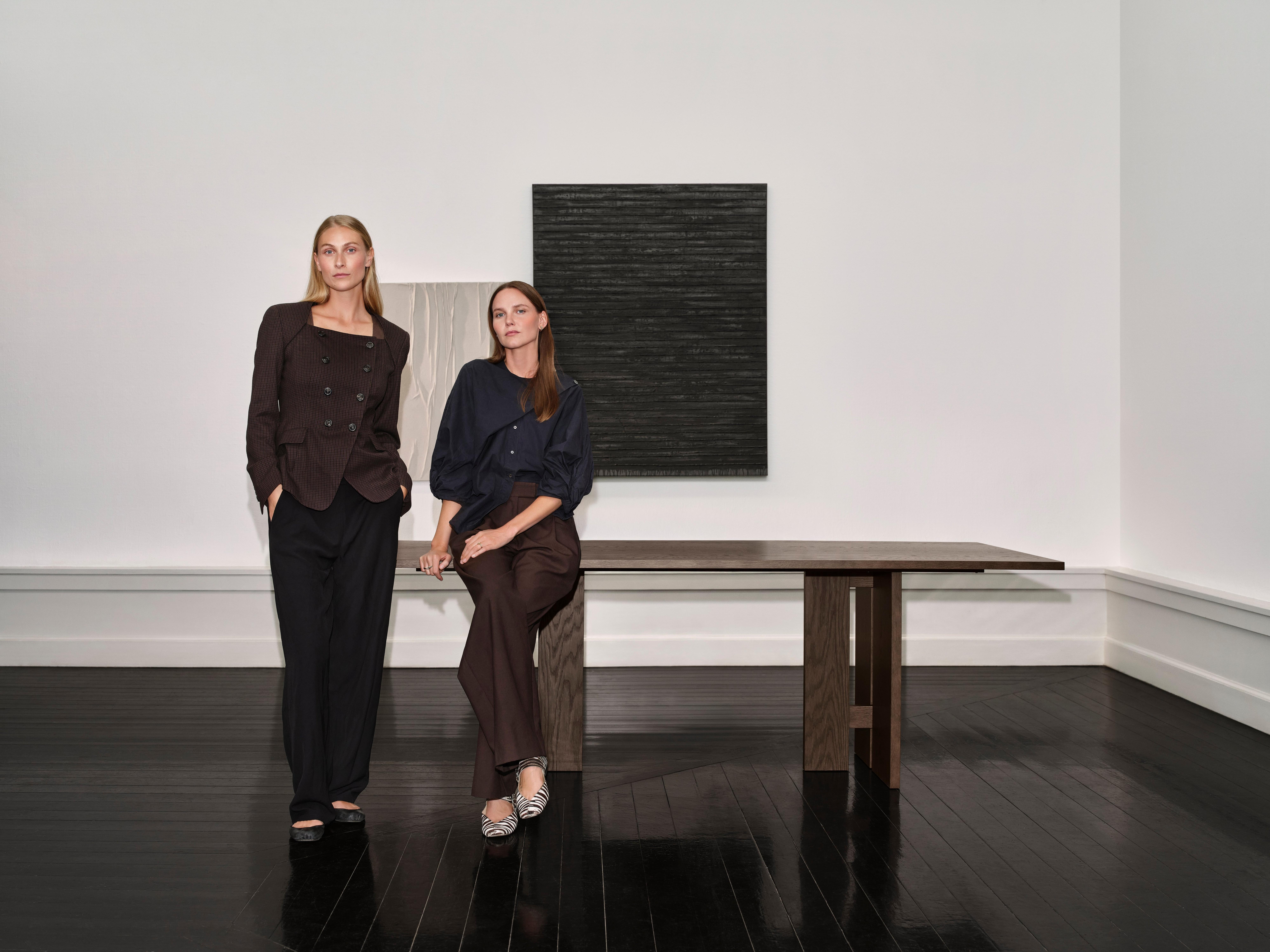 Two women in dark clothing pose by a wooden table. A textured artwork hangs on the wall.