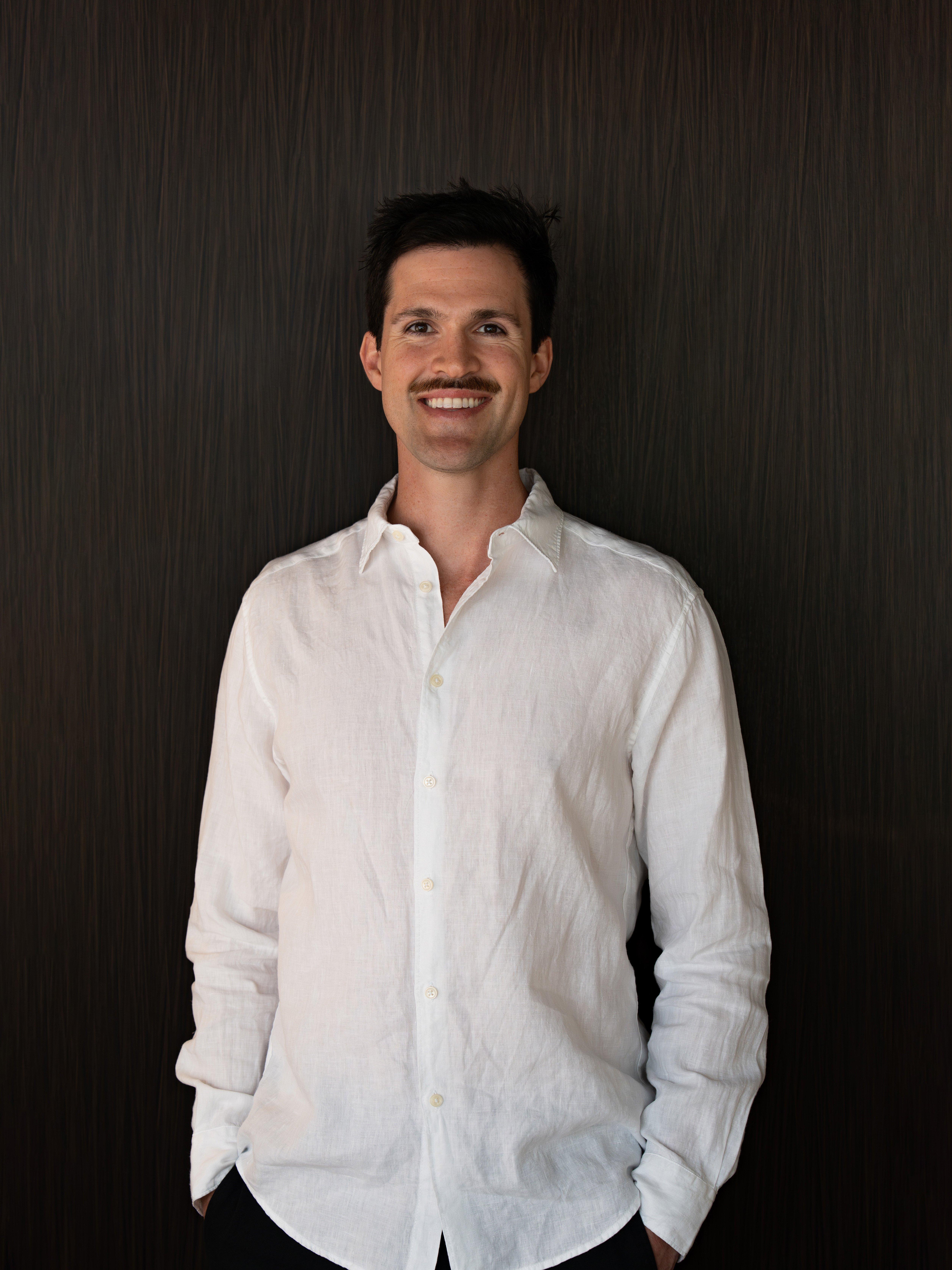 Model wearing a white long-sleeve button-up shirt, smiling with hands in pockets.