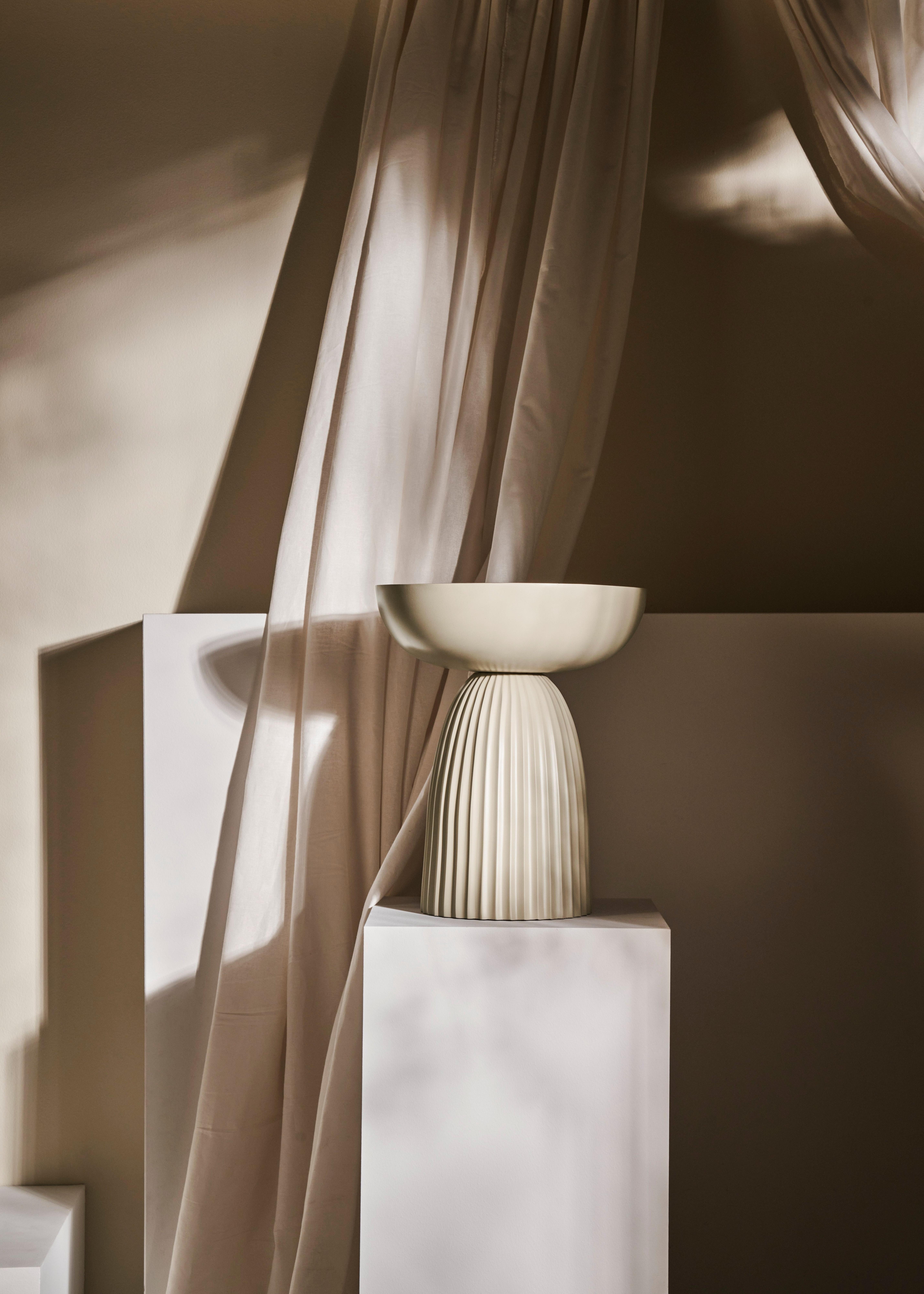 Élégante table d’appoint Expose gris chaud avec un bol plat sur le dessus, placée sur un fond de tissu beige fluide et de socles blancs, baignée d’une douce lumière naturelle.