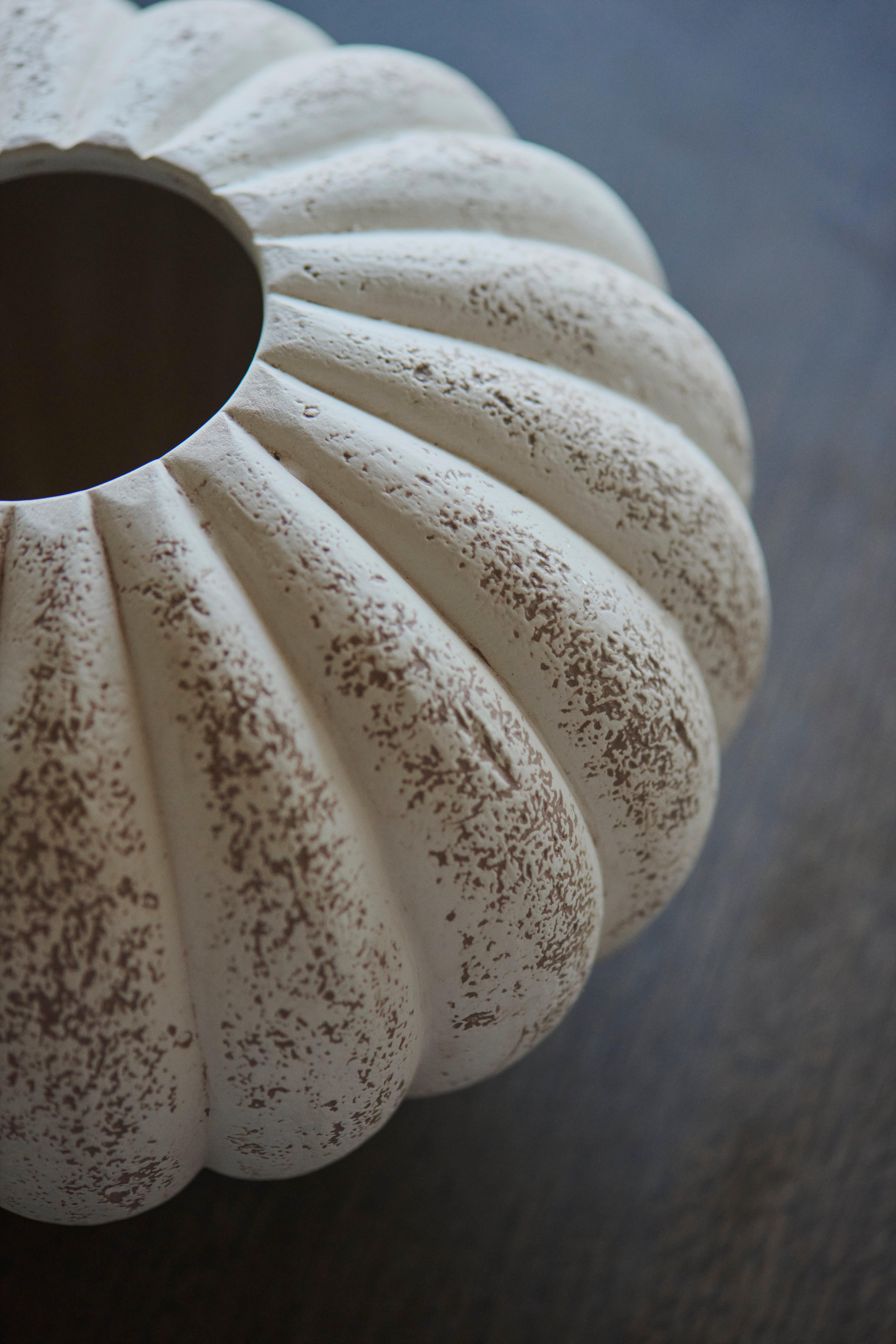 Nærbilde av en rund, keramikkvase med riller, patinert hvit finish og prikkete brun tekstur på en mørk overflate.