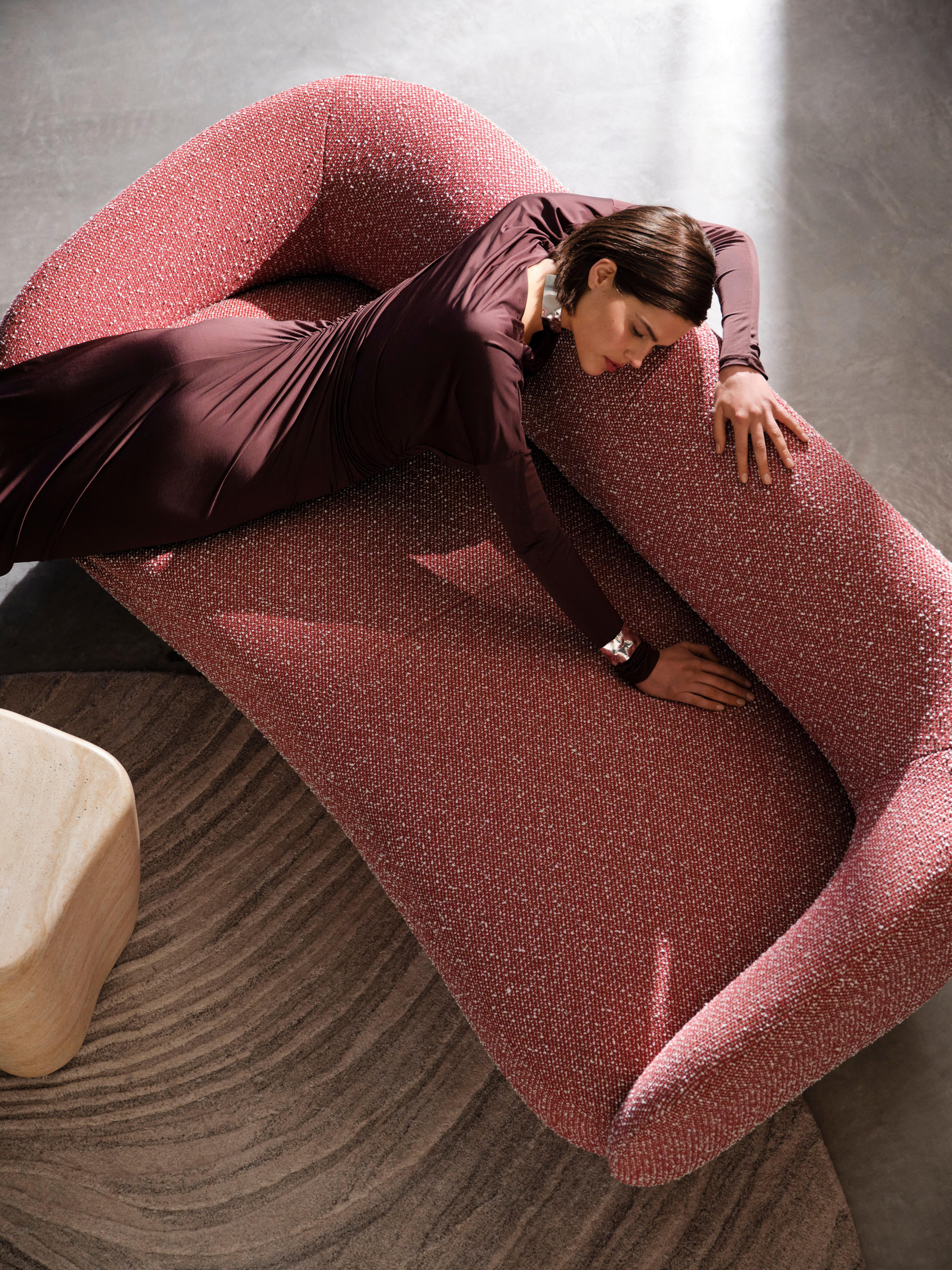 Mujer con un vestido granate descansando en un sofá Bolzano curvo rojo con una mesa auxiliar Terracina estilo travertino cerca de una alfombra gris texturizada.