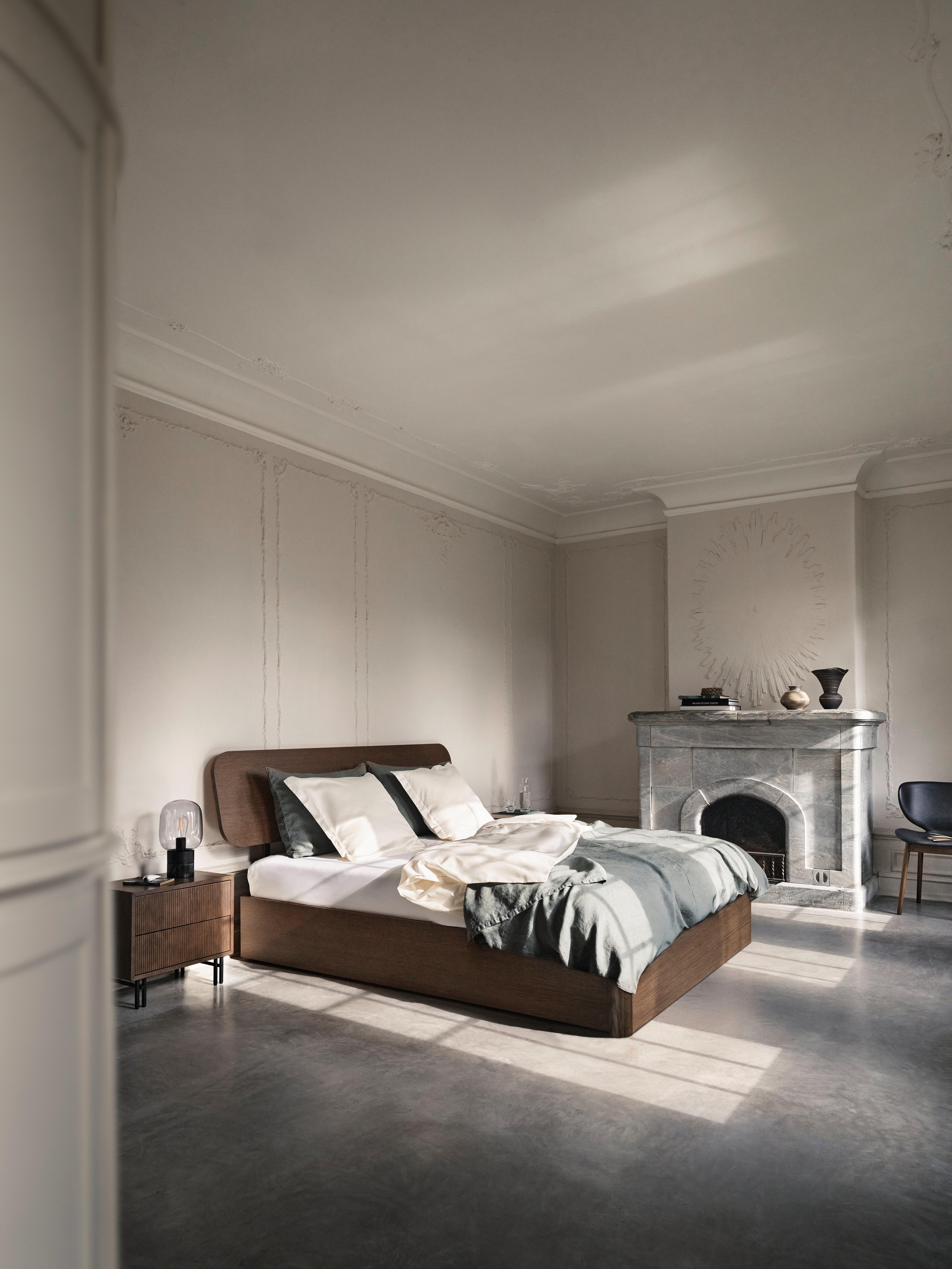 Minimalist bedroom with a low-profile bed, dark wood frame, grey bedding, side tables, and a marble fireplace.