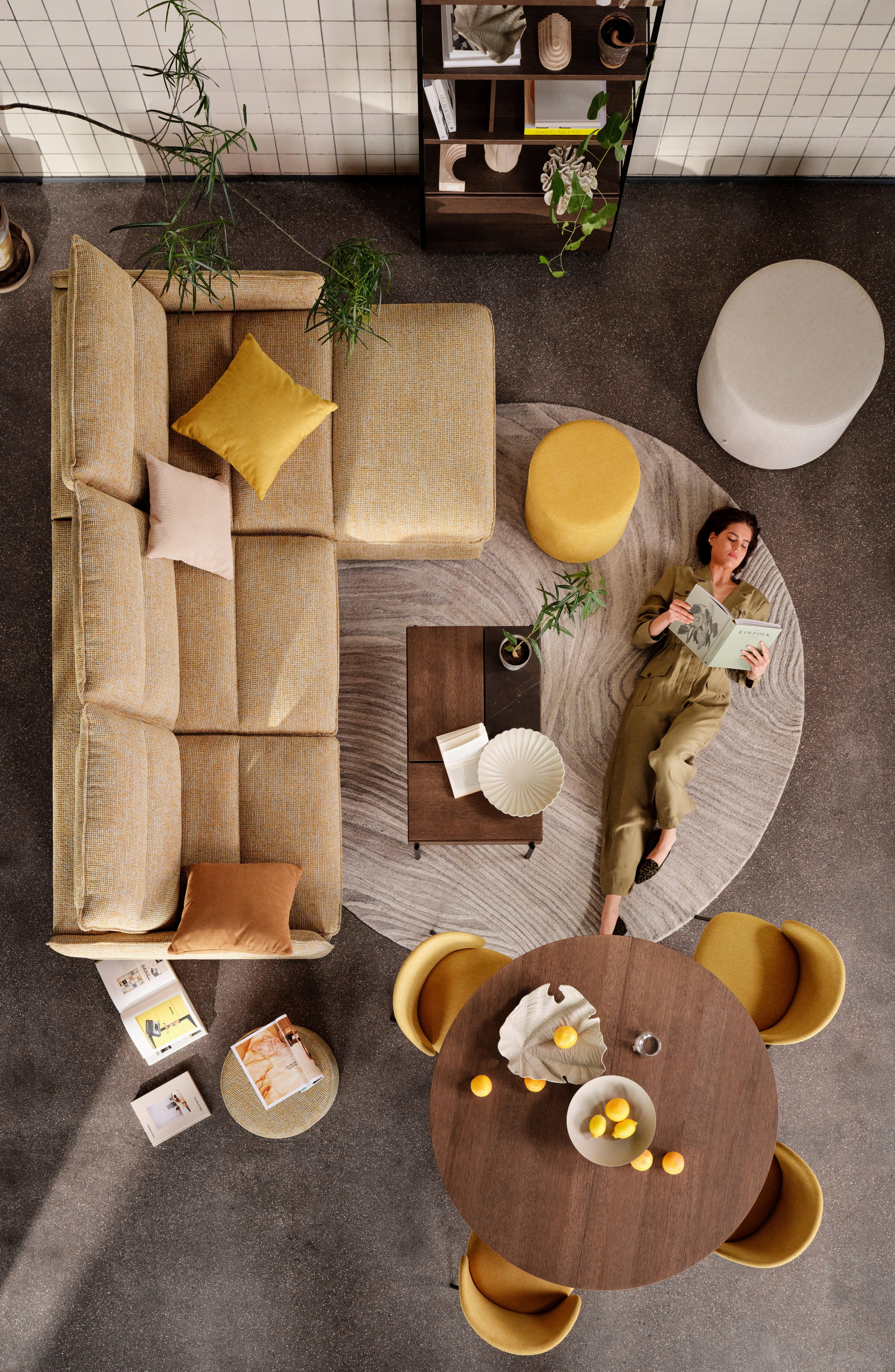 Whole room image of BoConcept furniture with model lying on the ground, featuring Southampton 2.5 seater sofa bed with resting unit, Chiva functional coffee table with storage, Santiago extendable dining table in dark oak veneer, Princeton chair in mustard Capri fabric, Eden footstool in mustard Capri fabric and various accessories