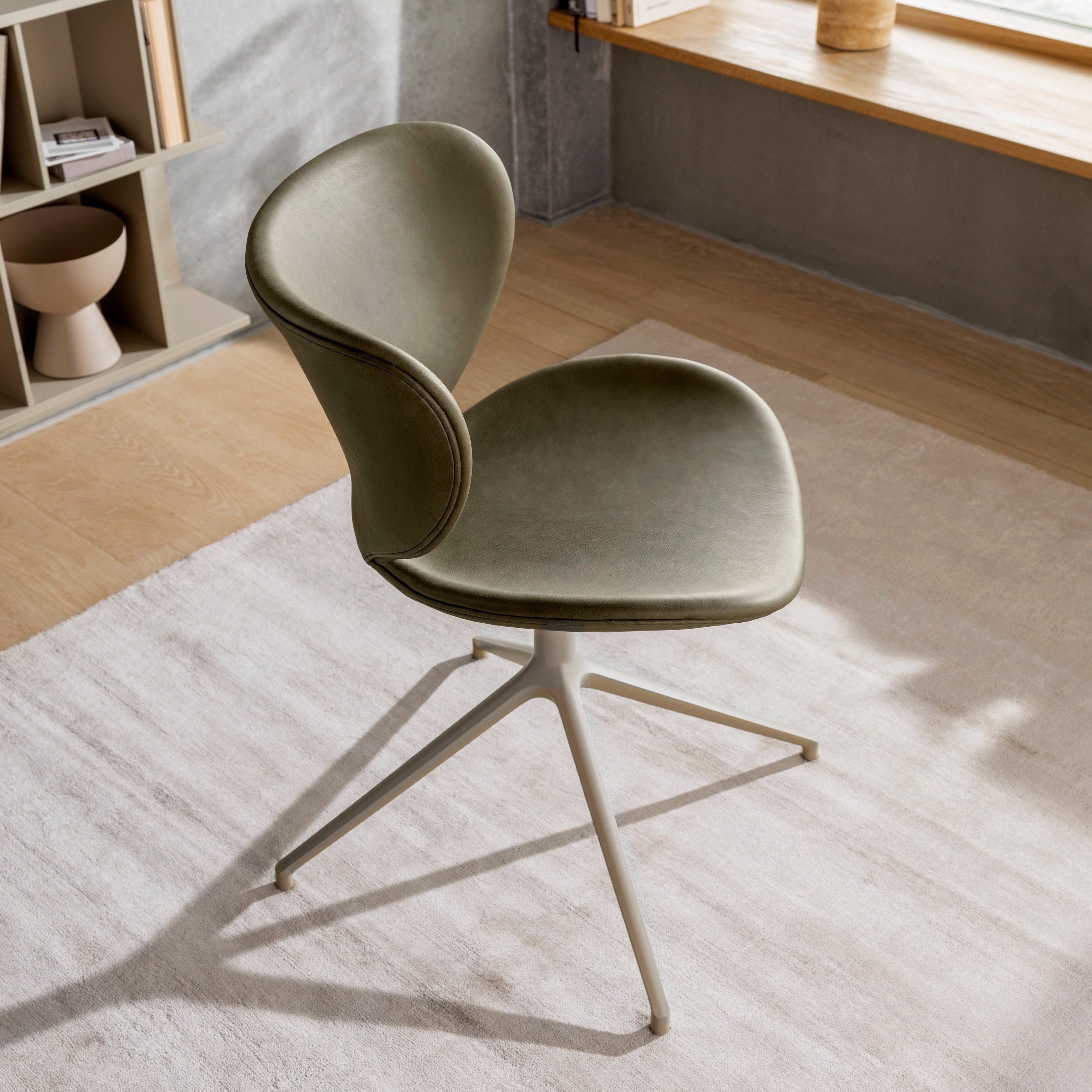 Olivově zelená židle s kovovou základnou v prosluněné místnosti s dřevěnými policemi a oknem.