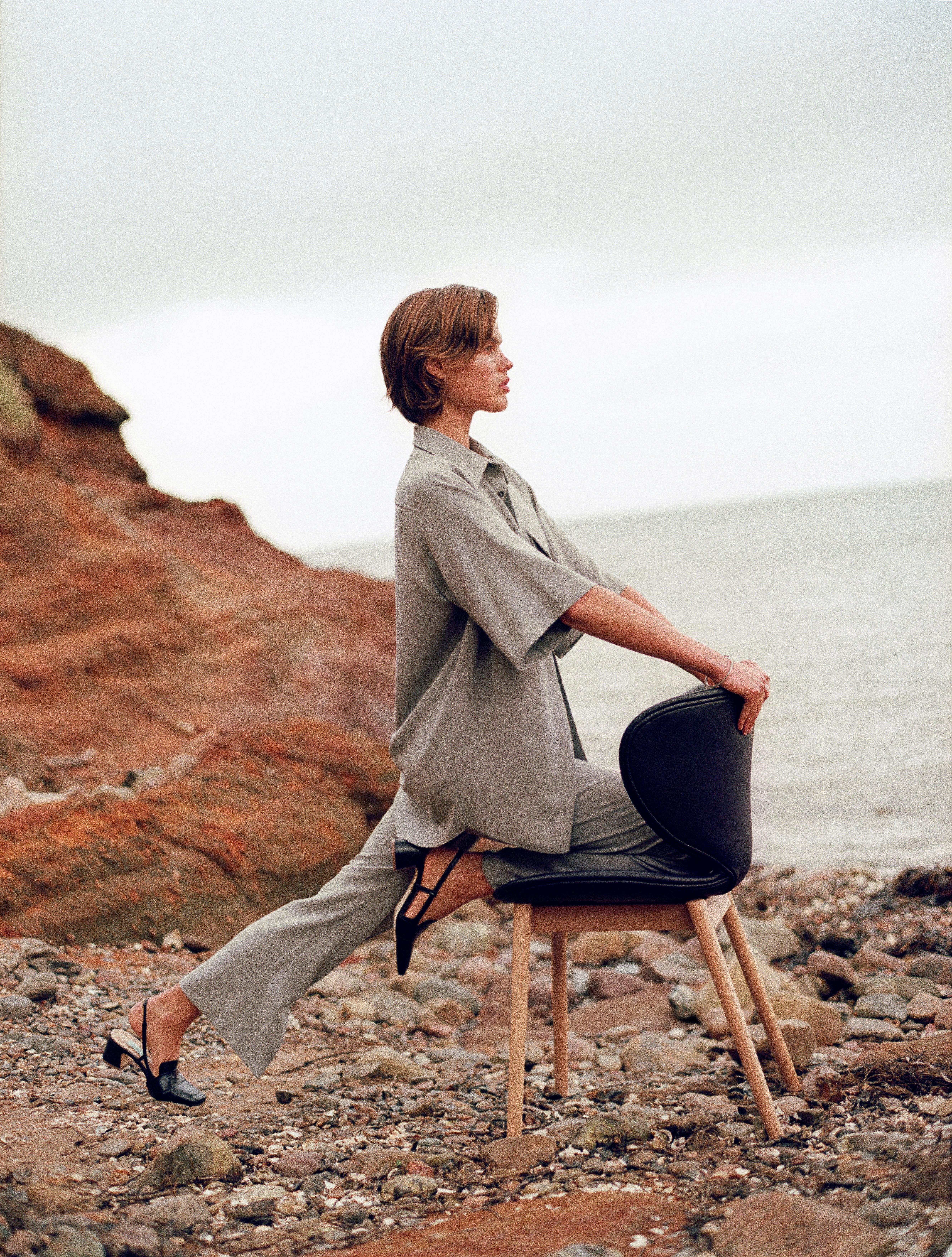 Woman in a grey outfit seated backwards on a Hamilton chair by a rocky shore, looking out to sea.