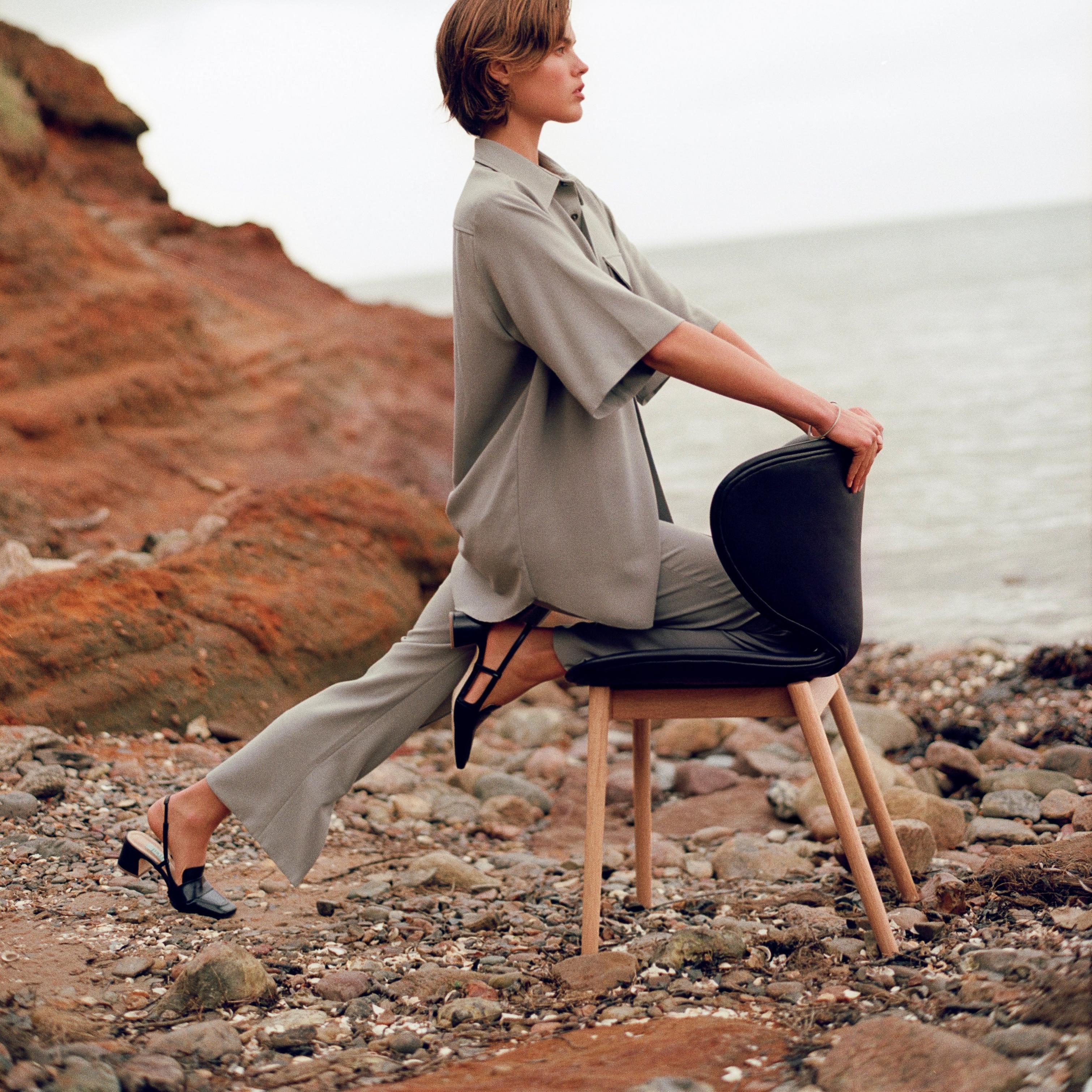Woman in a grey outfit seated backwards on a Hamilton chair by a rocky shore, looking out to sea.