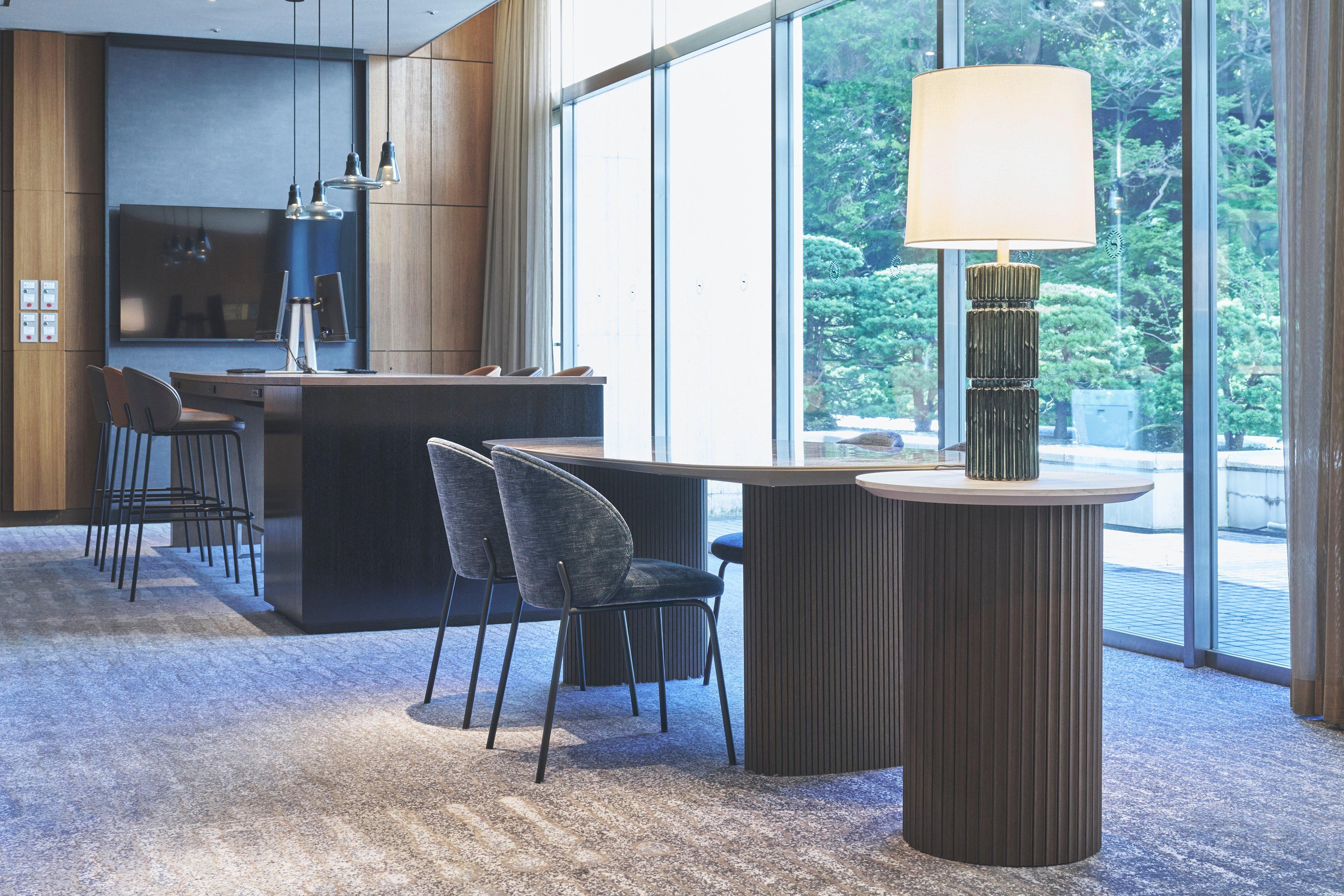 Modern lounge seating with tables, chairs, and a large table lamp by floor-to-ceiling windows.