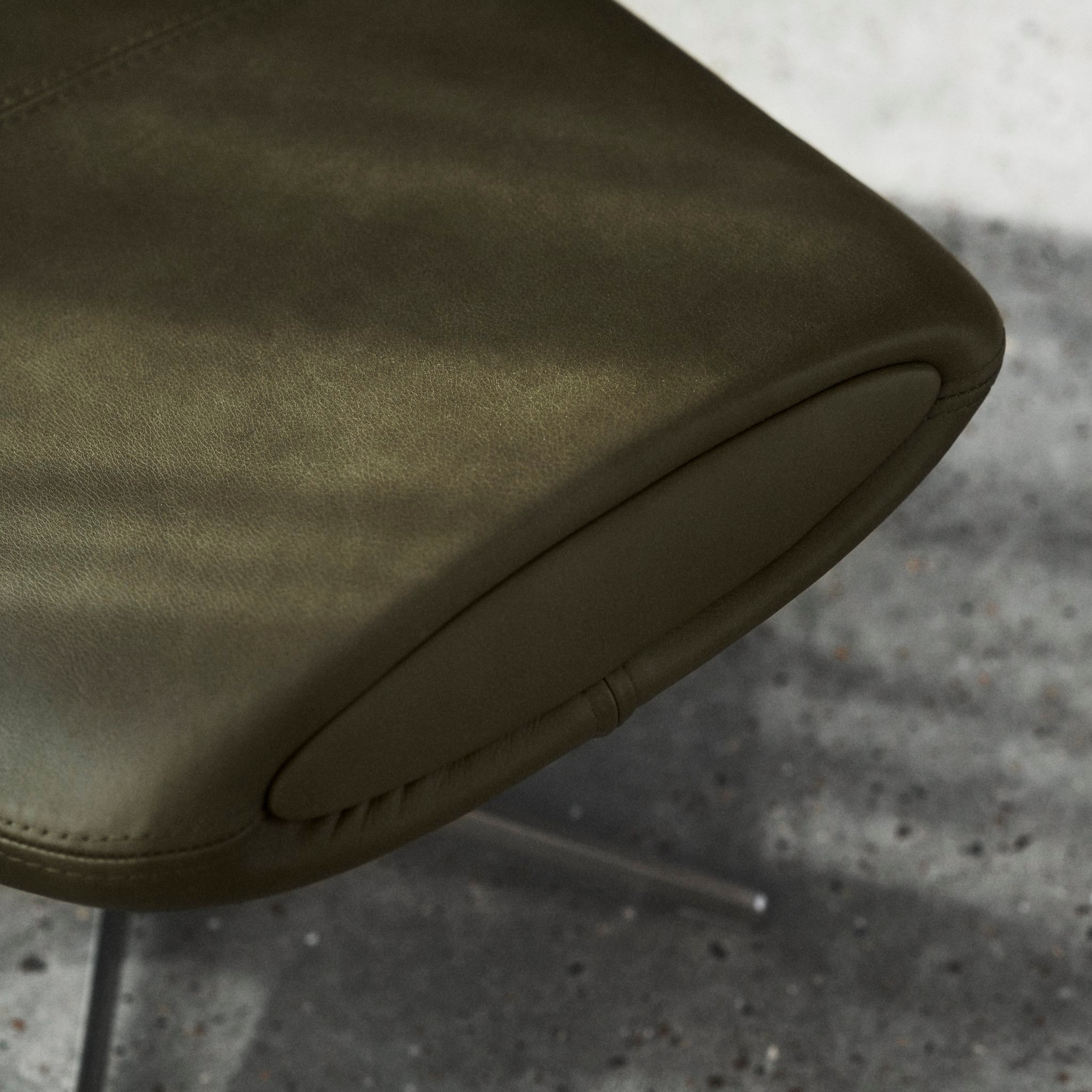 Nahaufnahme der Kante eines olivgrünen Stuhls mit Ziernähten auf einem Betonboden.