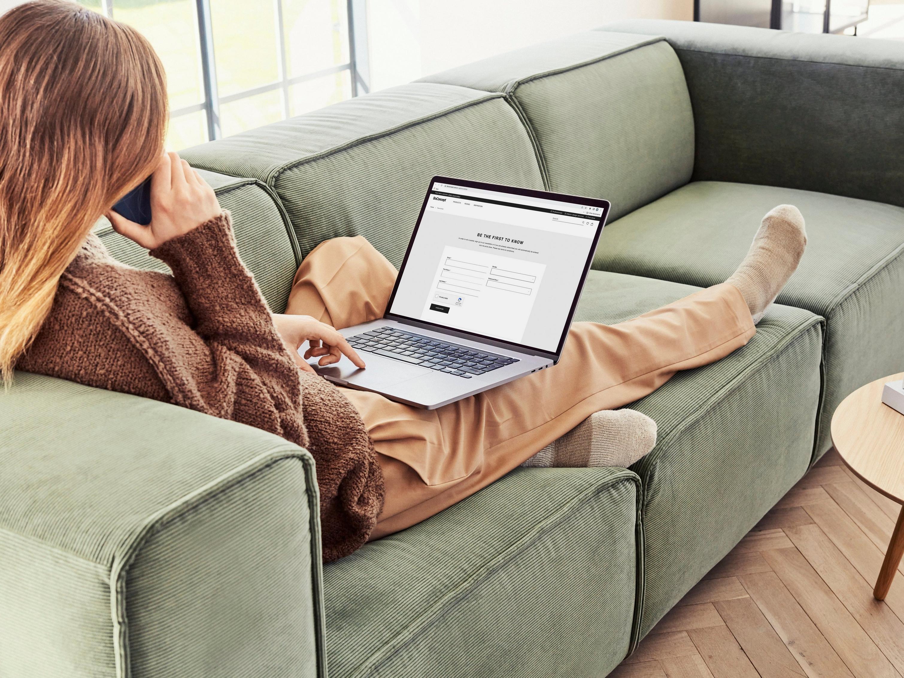 Person on a sofa using a laptop and phone, table with books nearby.