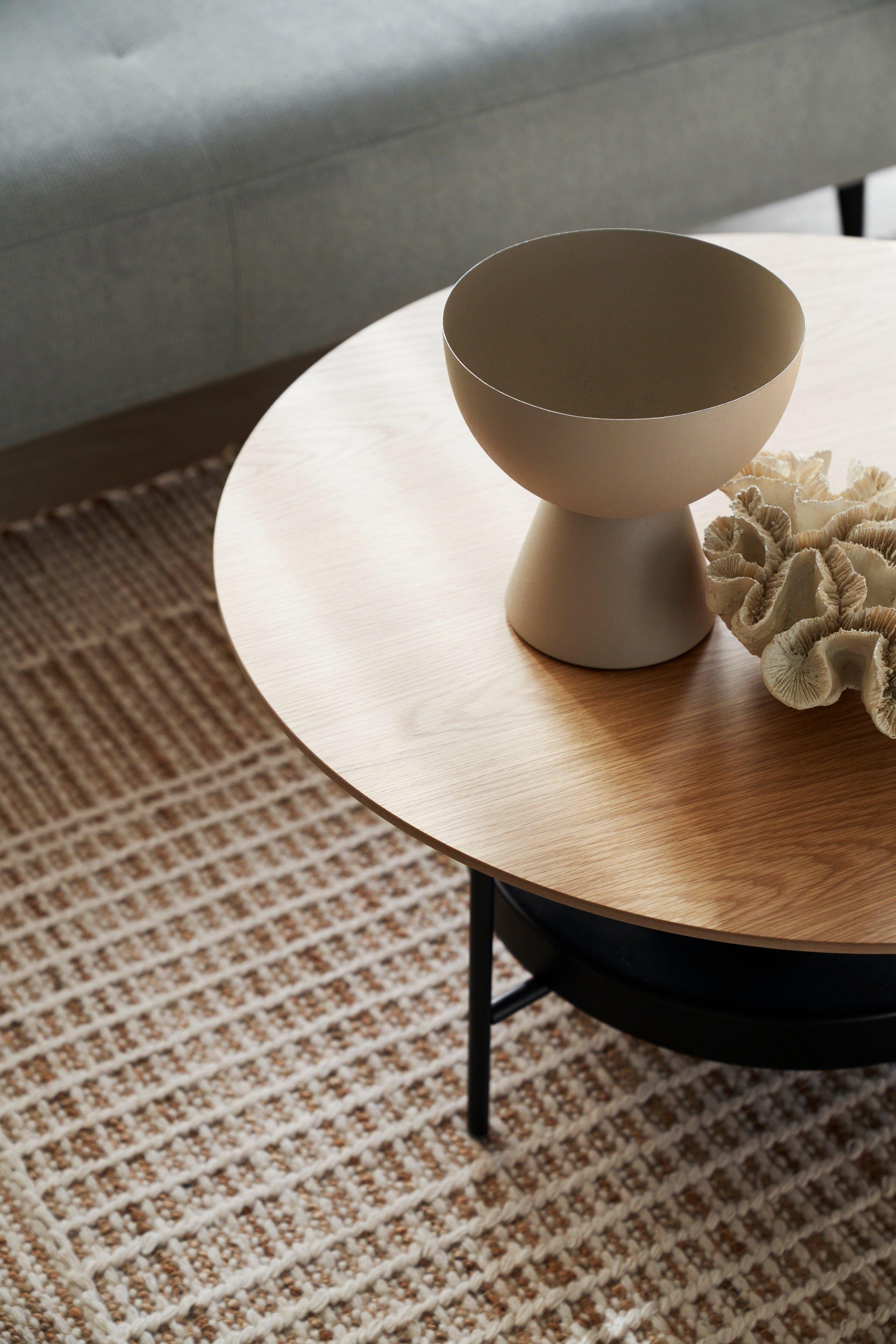 Round wooden Madrid coffee table with a vase and decorative item, near a sofa on a patterned rug.