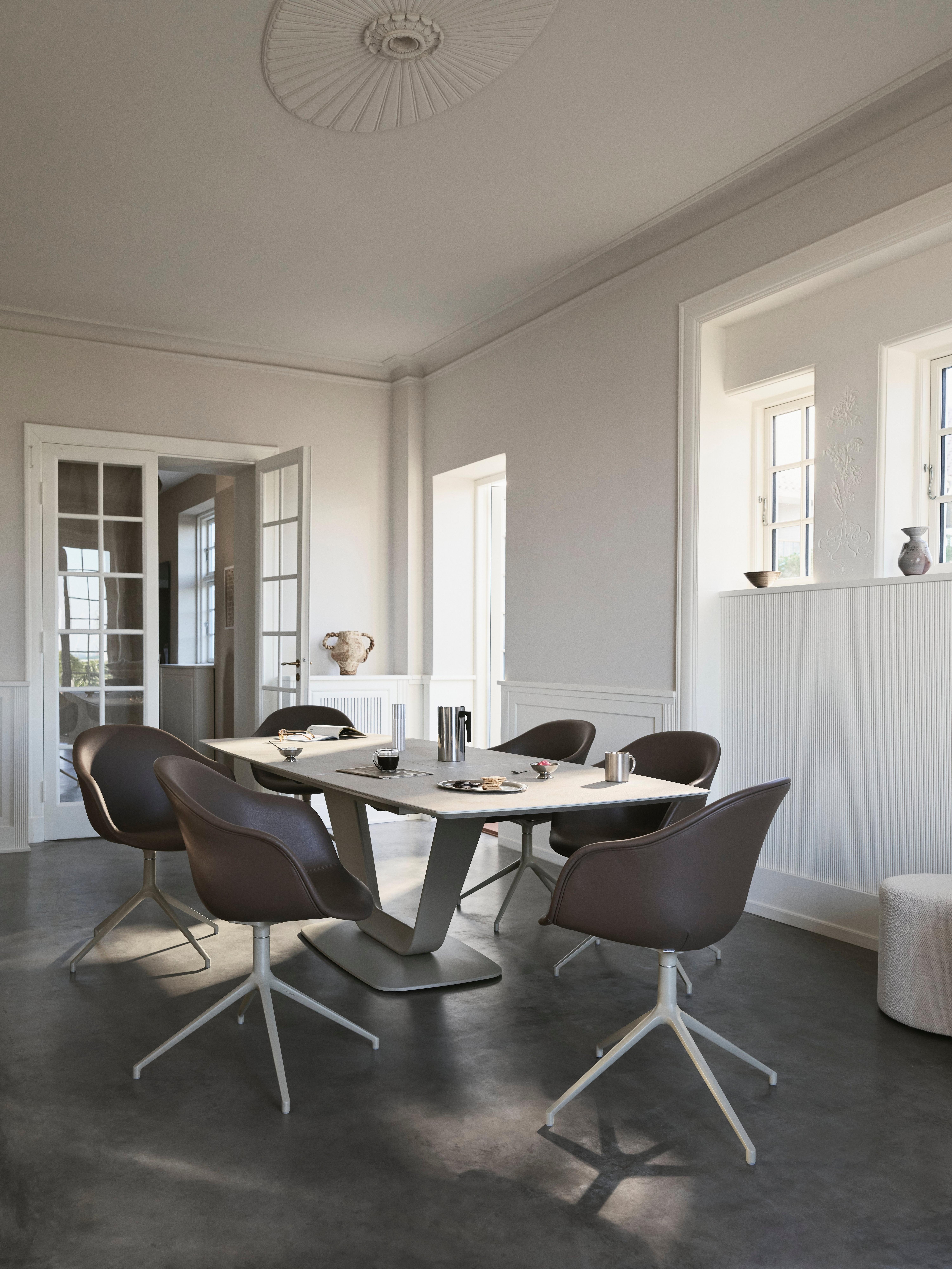 Modern dining room with six swivel chairs around a square table, set on a dark floor in a bright space with white walls and windows.
