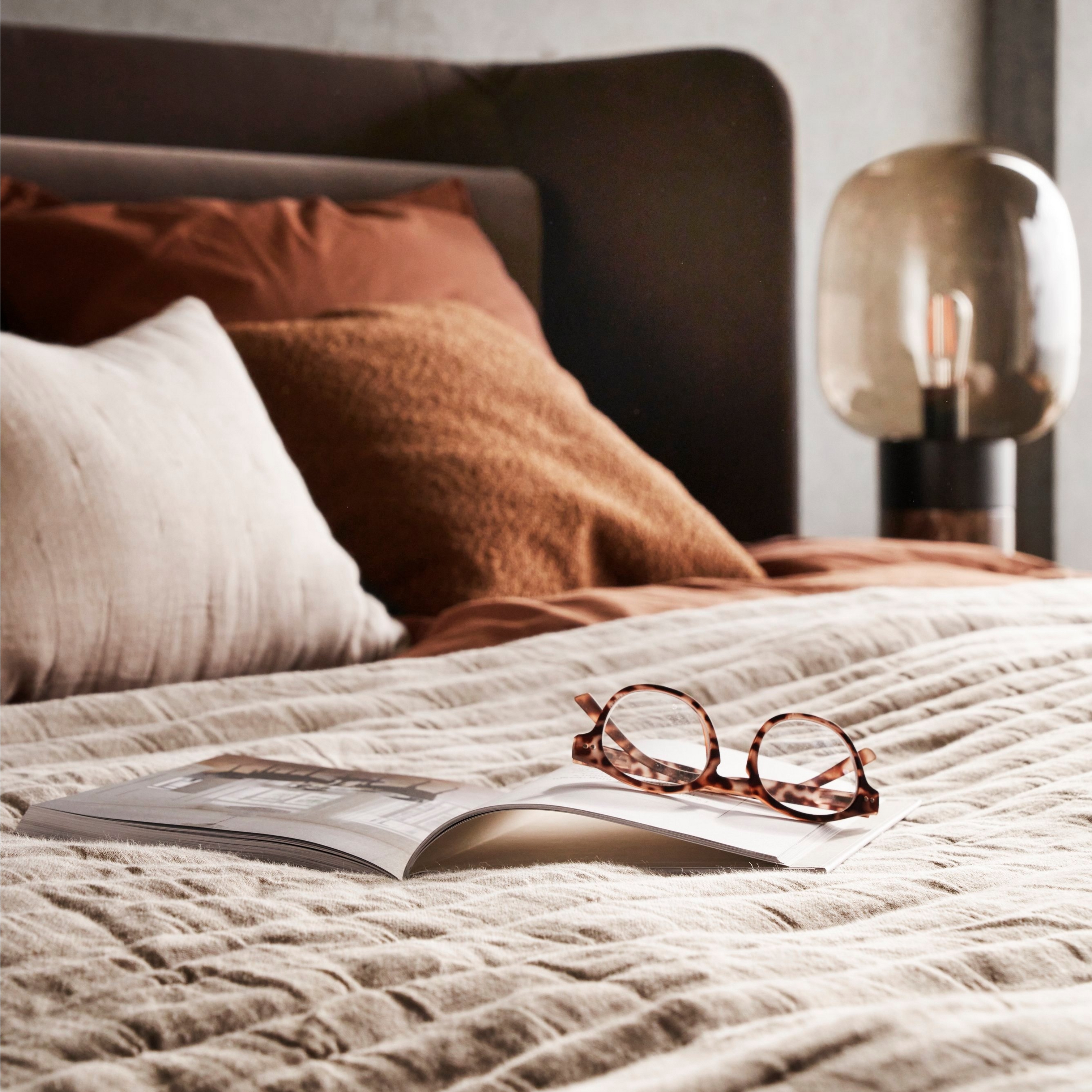 Cama com roupa de cama texturizada, óculos pousados num livro aberto e iluminação suave na mesa de cabeceira.