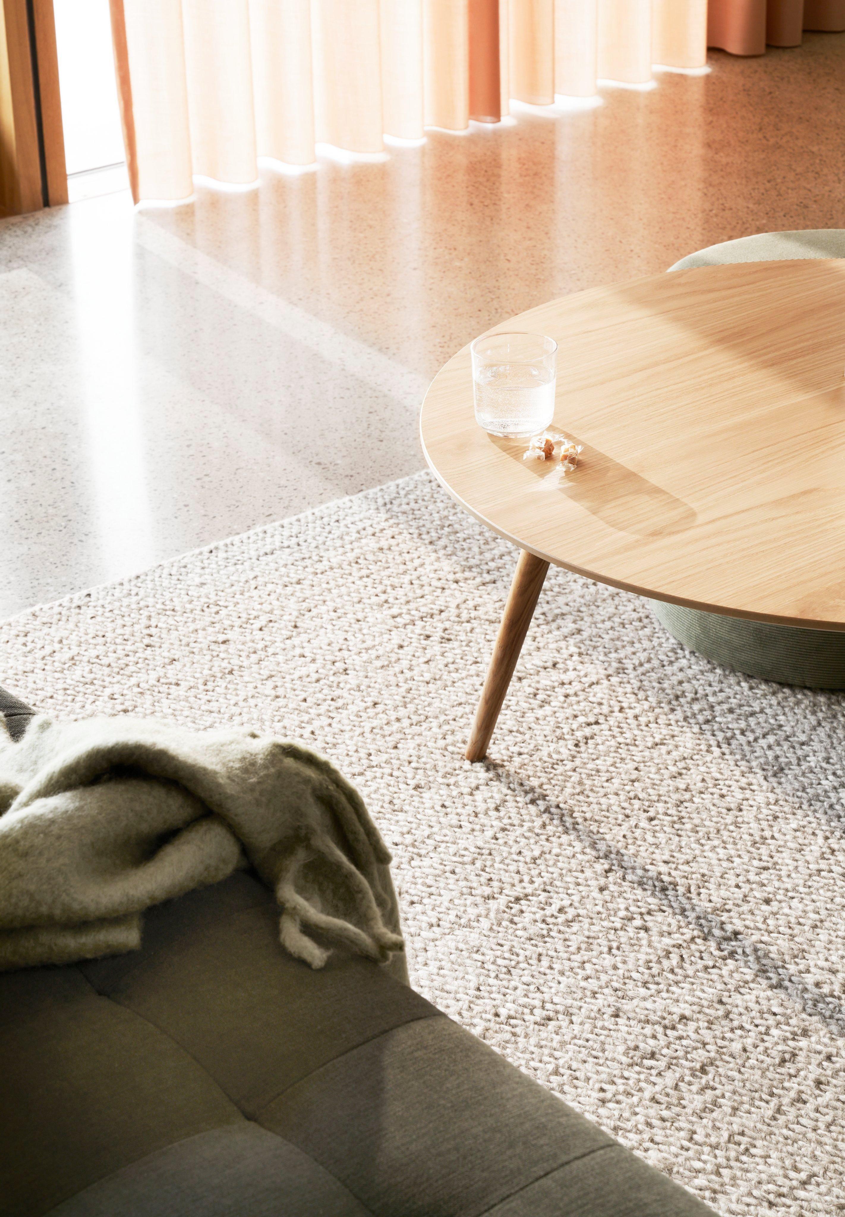Kuvioitu matto ja pyöreä puupintainen pöytä, lasillinen vettä ja pehmeä huopa auringonvalossa.