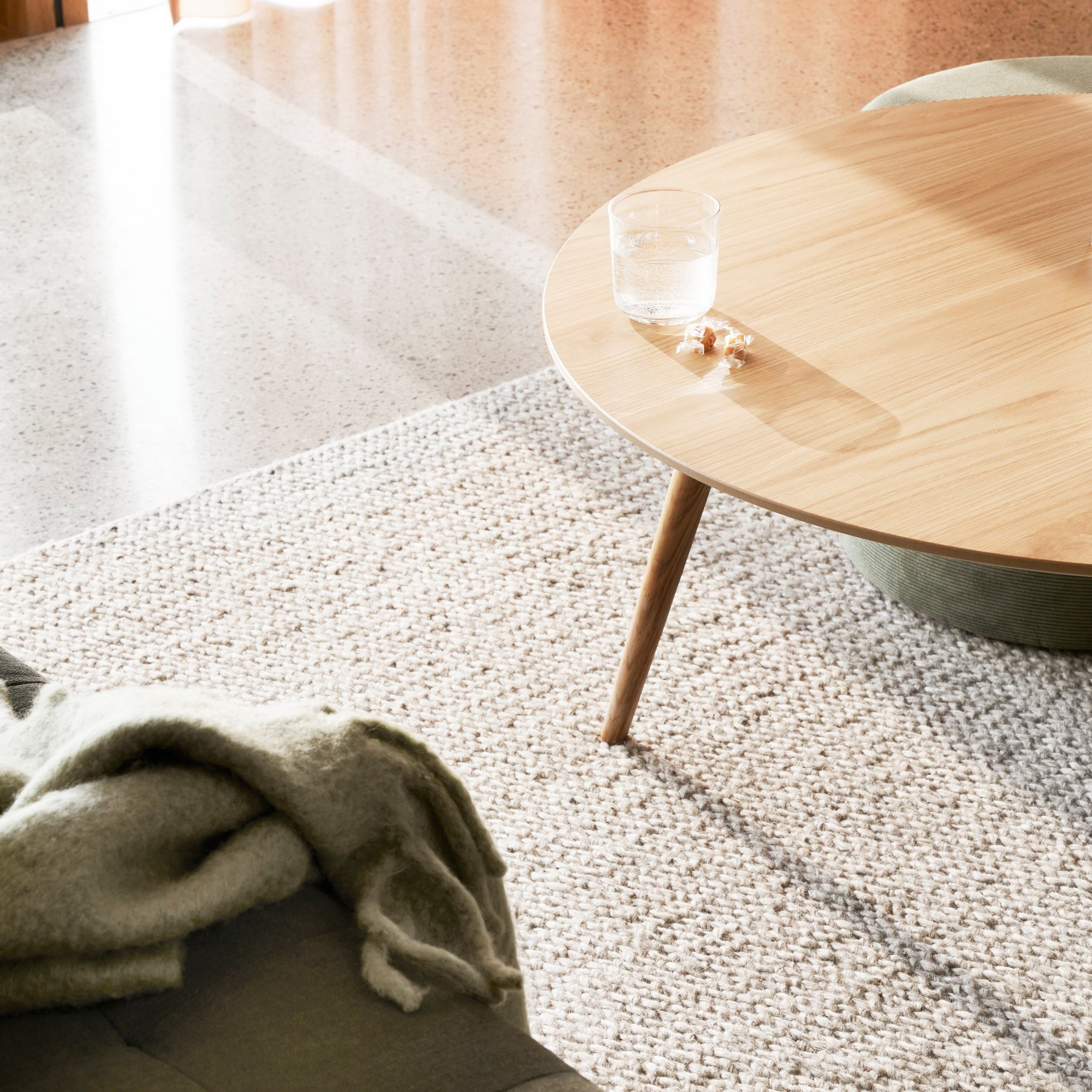 Textúrovaný koberec s okrúhlym dreveným stolom, pohárom vody a mäkkou dekou na slnečnom svetle.