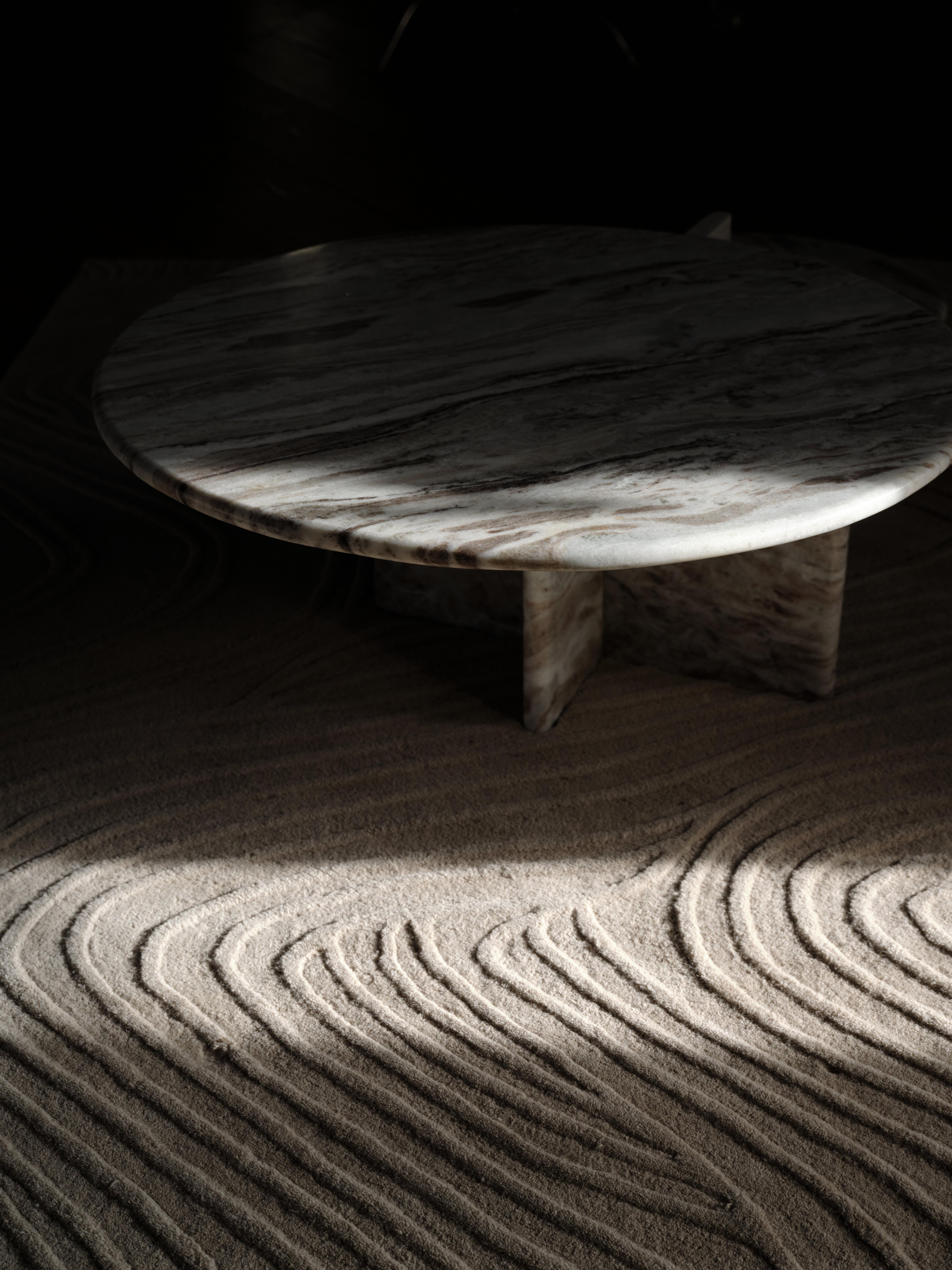 Round marble coffee table on textured sand-colored rug with sculpted wave-like patterns, lit by dramatic natural light.