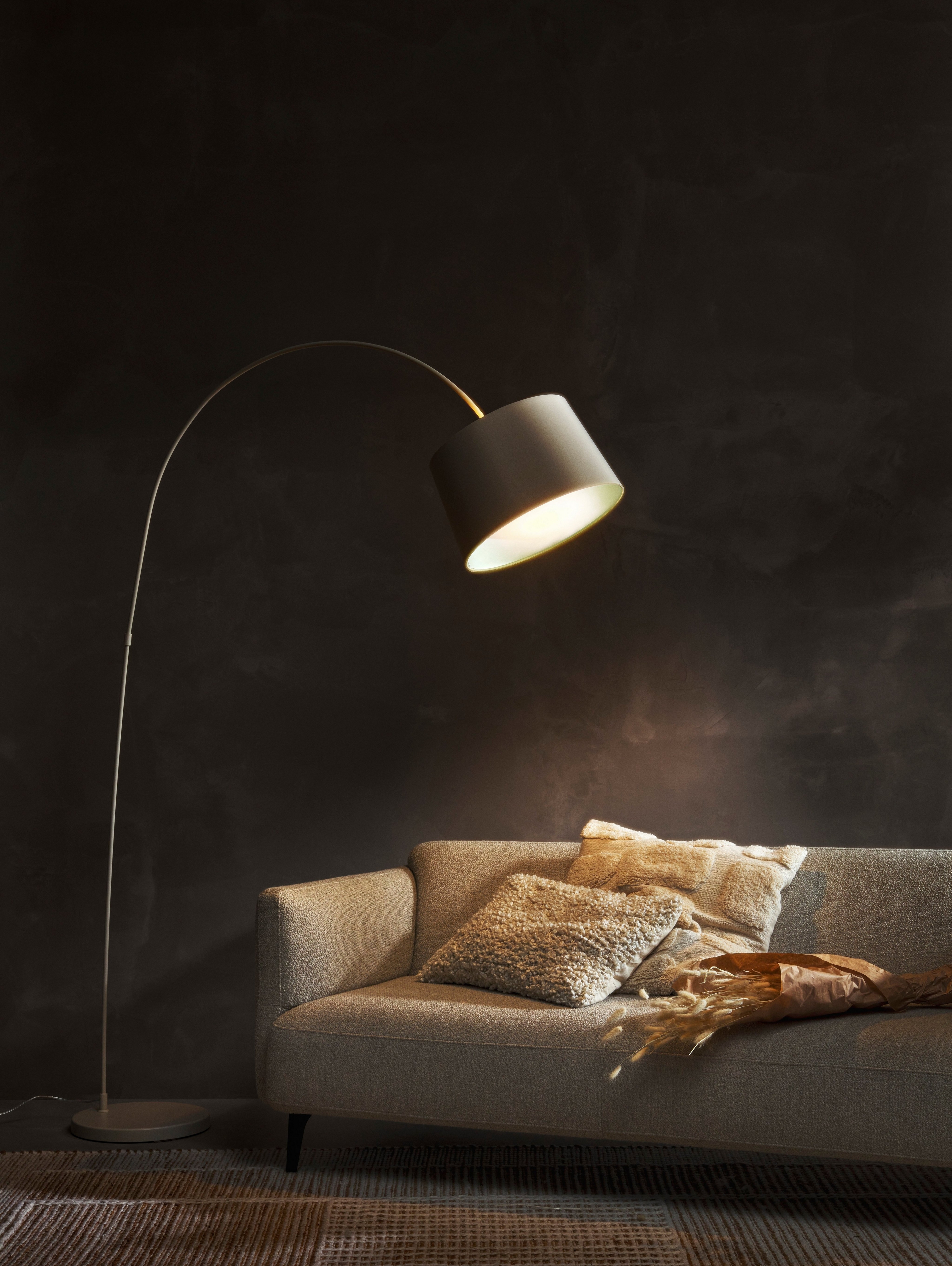 Candeeiro de chão Kuta a iluminar um sofá Modena cinzento com almofadas e um fundo de parede escuro.