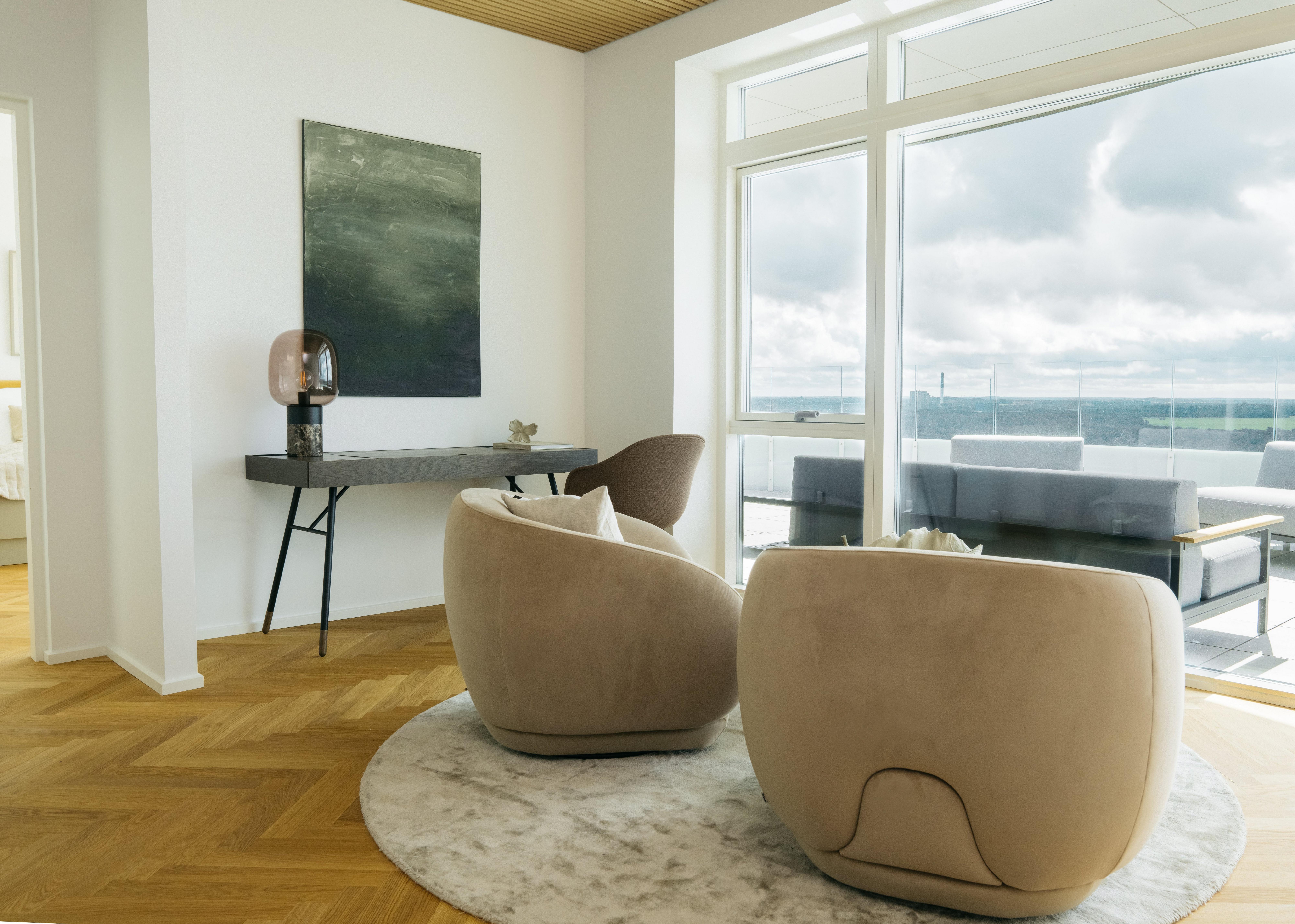 Seating area with two beige rounded chairs on a circular rug, a black console table, and a large window overlooking a balcony with outdoor seating.
