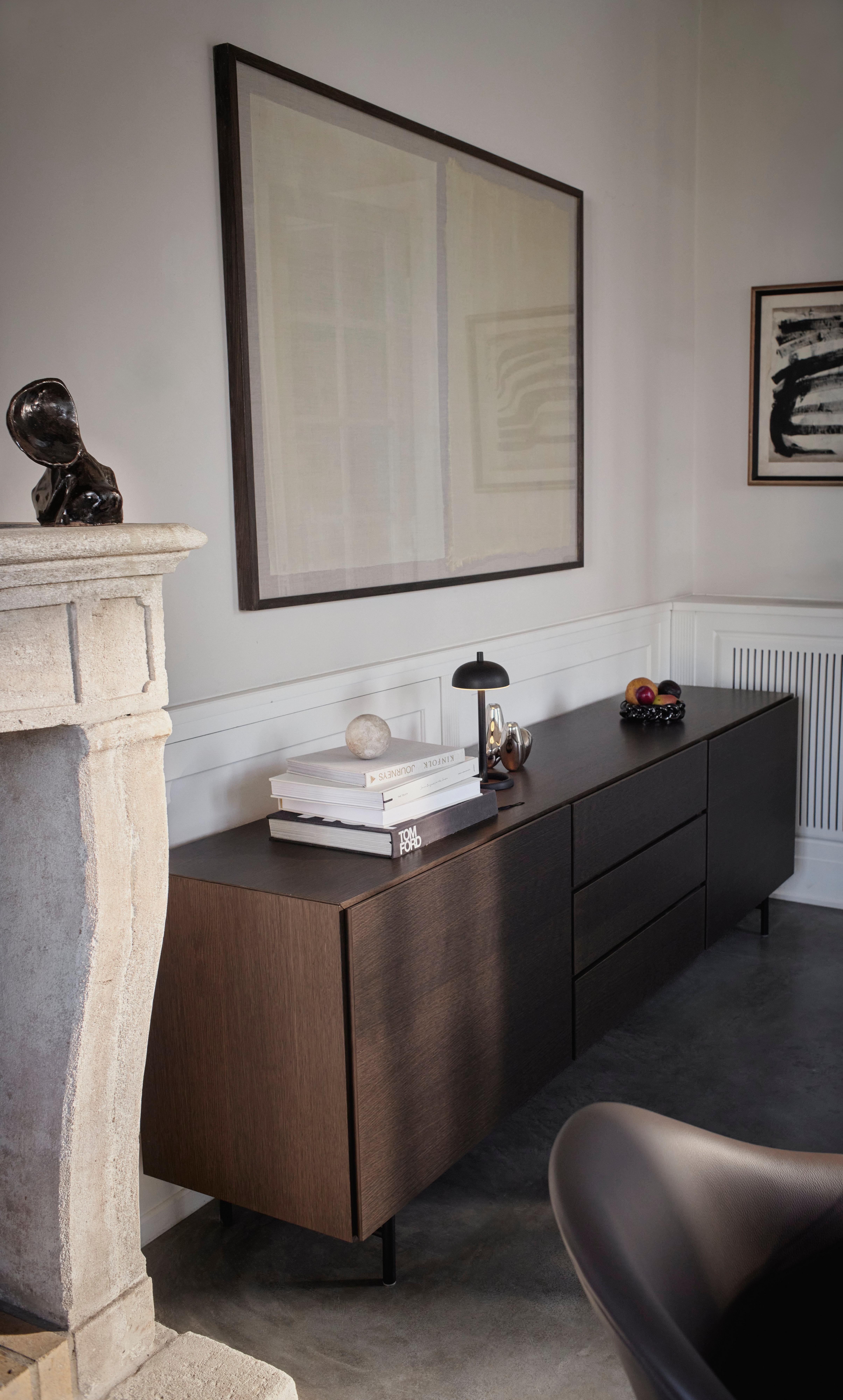 Dark wood sideboard with books and sculptural decor, beside a stone fireplace and abstract framed artwork on pale walls
