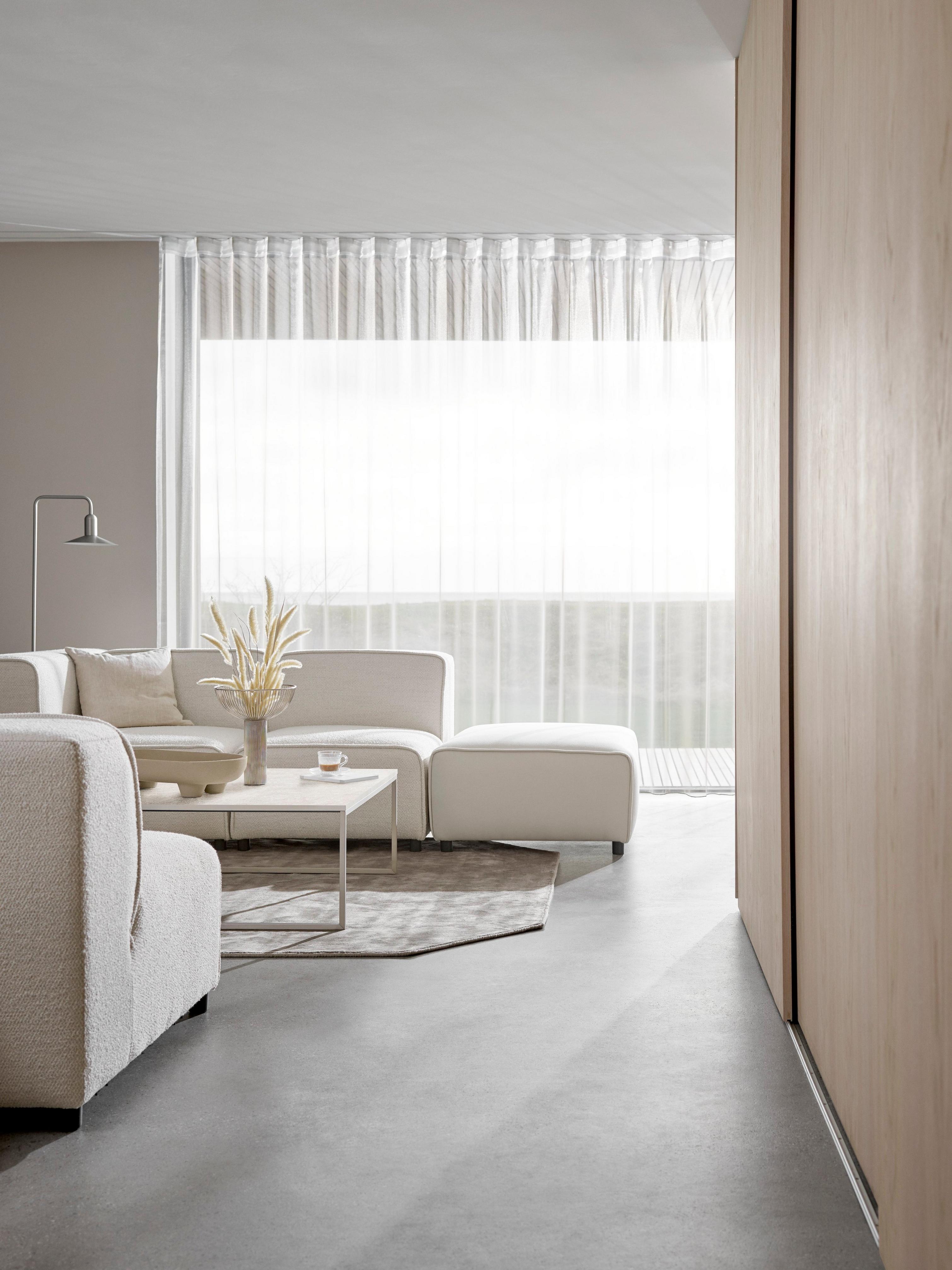 Cremefarvet Carmo sofa i minimalistisk stue og transparente gardiner, der spreder lyset.