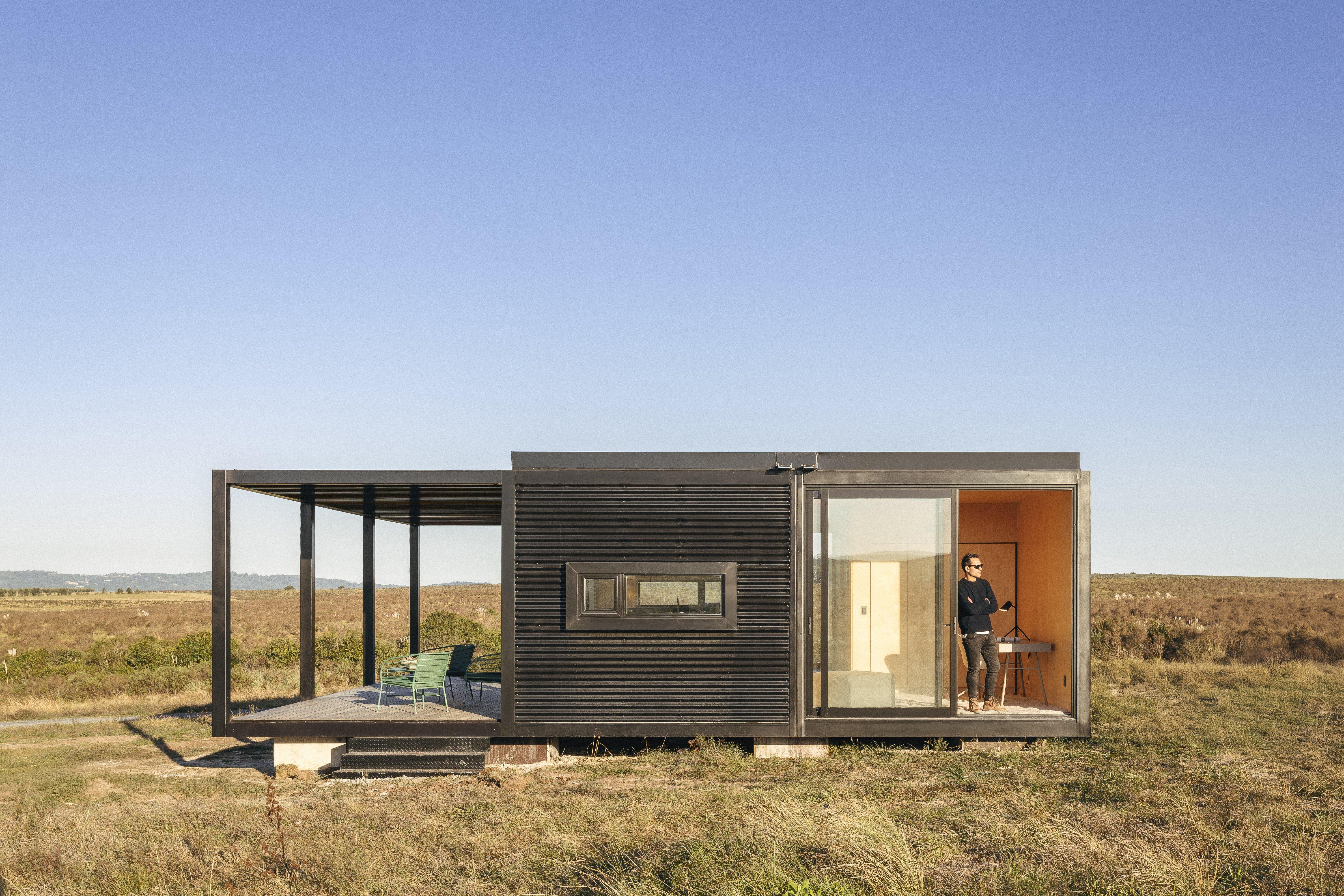 現代化的小屋在草地上，一人站在玻璃門旁，外有露台和綠色椅子。