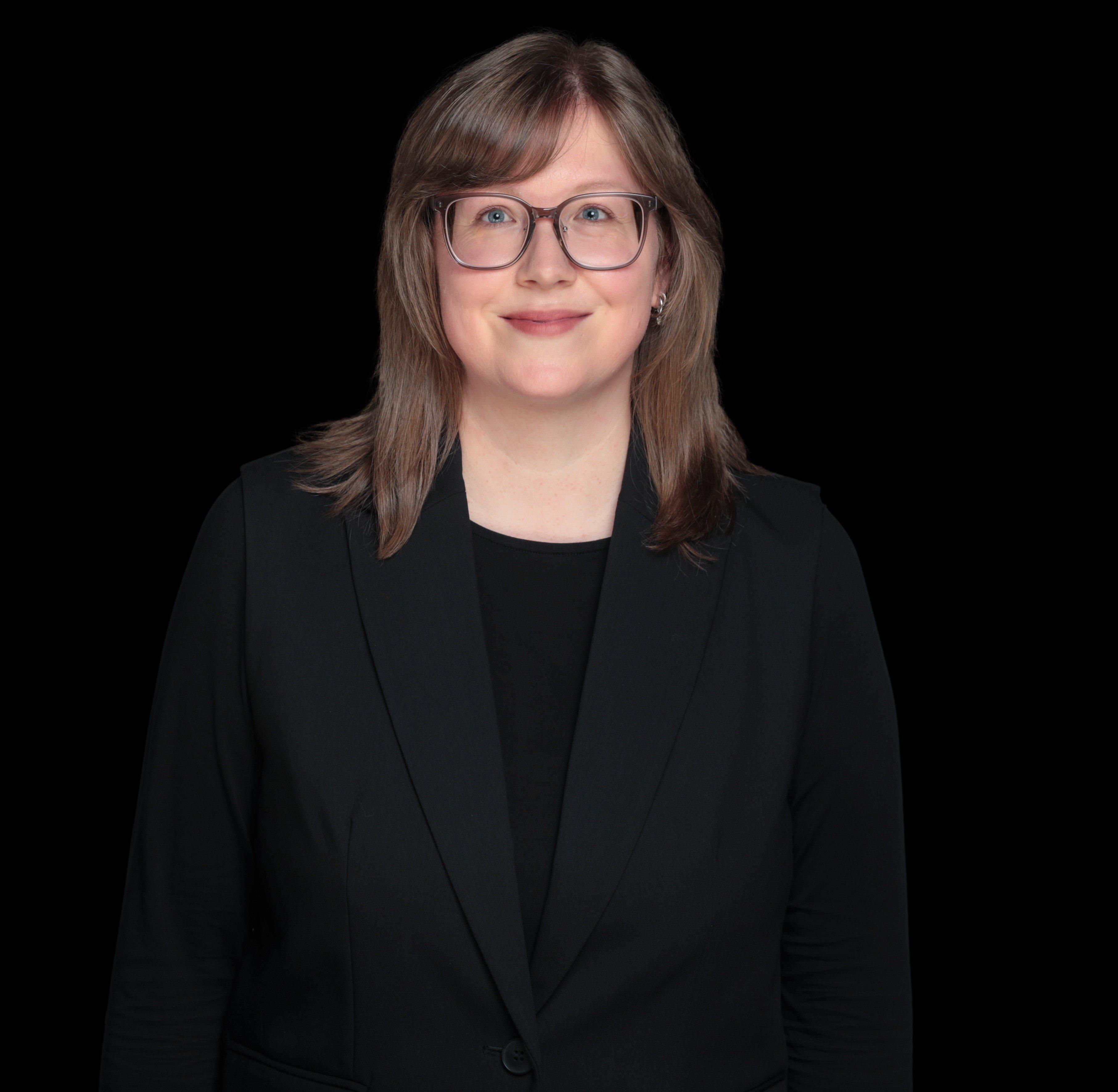 Porträt einer Frau mit Brille im schwarzen Blazer, lächelt in die Kamera.