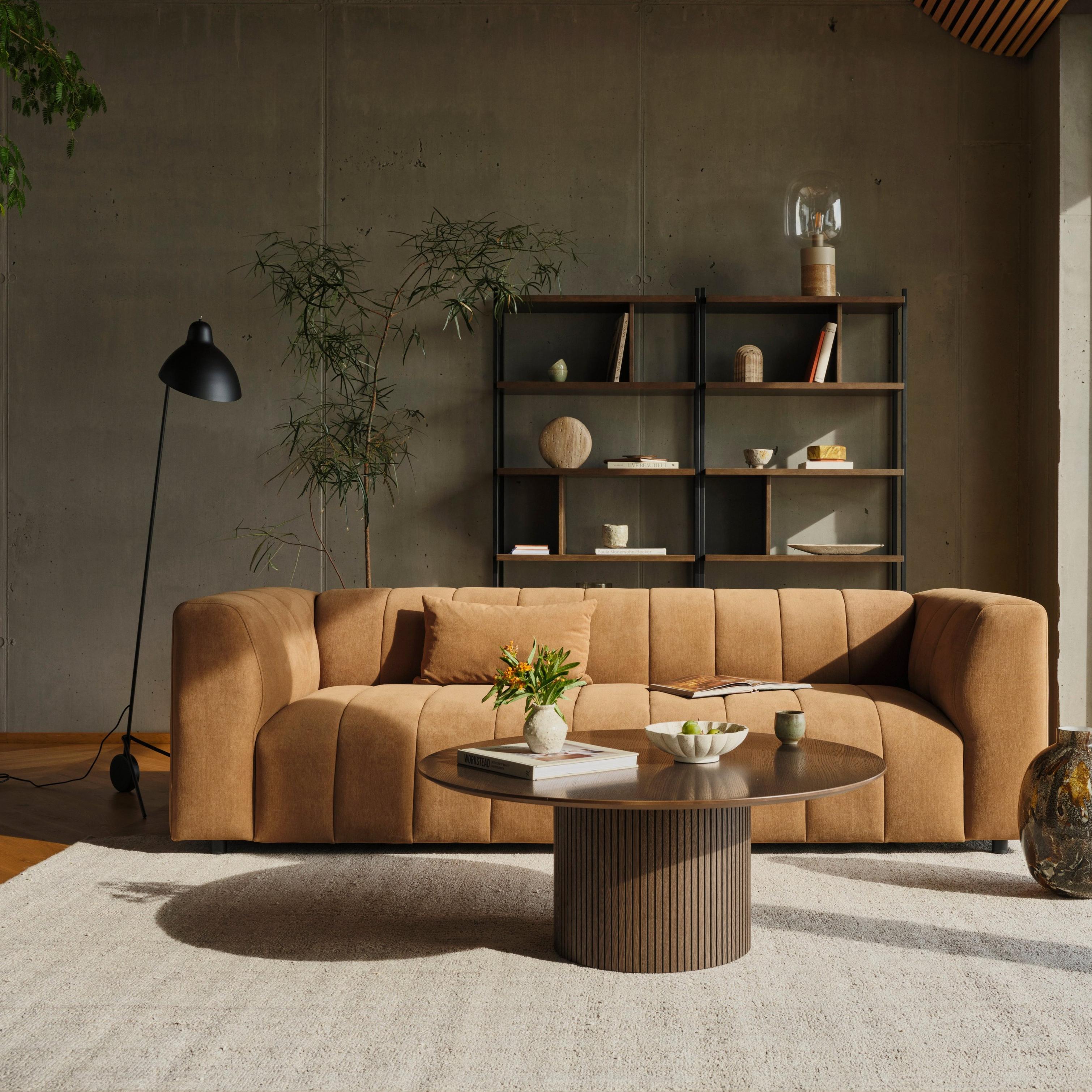 Stylish living room with a tan sofa, round table, floor lamp, and bookshelf by a large window.